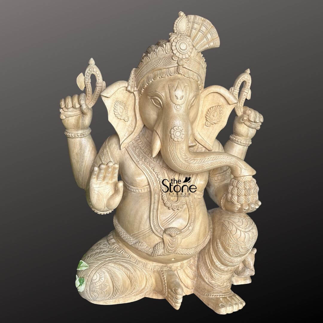 Wooden Ganesha Statue Online 2ft 2 A highly decorative 2-foot wooden Ganesha statue is presented from a frontal slightly elevated perspective against a plain dark background highlighting its intricate details. The statue is meticulously carved from light-colored wood showcasing its natural grain and warm inviting tones which would suit various home decor styles. Lord Ganesha is depicted in a traditional seated posture with one knee raised exuding an aura of wisdom prosperity and benevolence. Ganesha is shown with four arms his upper hands hold traditional attributes, possibly a noose and an axe. His lower right hand is raised in a gesture of blessing offering protection, while his lower left hand rests on his lap possibly holding a modak or sweet dumpling. His elephant head is intricately detailed with large, fan-like ears, expressive eyes, and his trunk curving gracefully towards his left. He wears an elaborate crown adorned with a distinctive fan-shaped element at the back, along with intricate necklaces, armlets, and a sacred thread across his plump belly. His garments are finely sculpted with delicate folds and patterns, enhancing the traditional and decorative aesthetic. A small mouse Ganesha's vehicle is subtly carved near his foot, though partially obscured. The natural wood finish adds to its rustic and handcrafted charm. This detailed image vividly conveys the spiritual majesty and artistic craftsmanship of the wooden Ganesha statue making it highly relevant to an article discussing decorative wooden Ganesha idols.