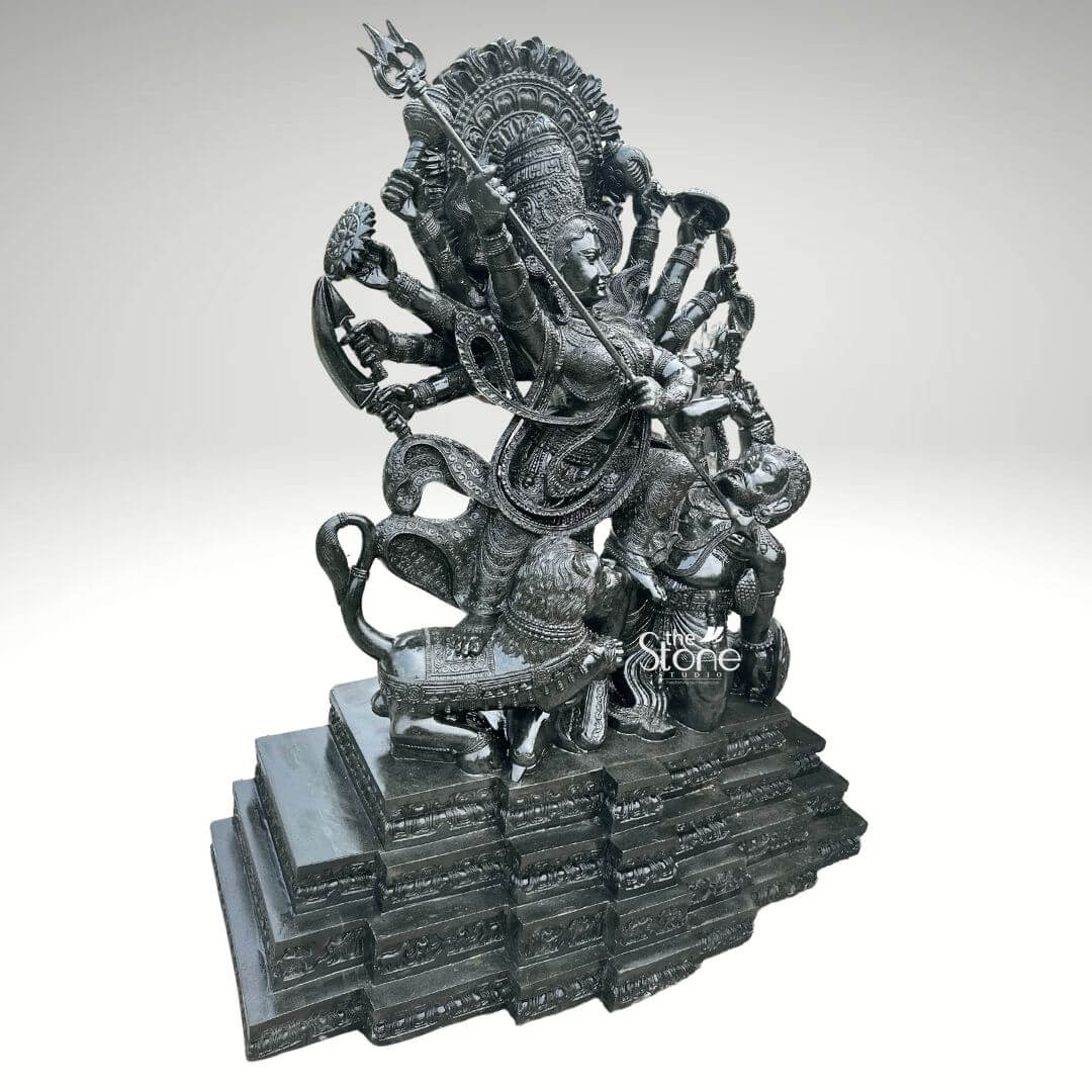 6ft Mahishasura Mardini Durga Statue 12 A majestic 6-foot tall Durga Mahishasura Mardini statue masterfully hand-carved from premium natural black stone serves as a grand spiritual centerpiece for a temple foyer or a luxury outdoor landscape. This monumental 6ft blackstone Durga idol captures the Goddess in her powerful ten-armed form, depicting the iconic moment of divine triumph as she slays the buffalo demon, Mahishasura. Seen from a three-quarter side perspective to highlight its monumental scale and intricate depth, the deity is shown in a dynamic warrior pose. Her central hands are shown thrusting a long, slender trident (trishula) into the demon, who is depicted in human form emerging from the buffalo. Goddess Durga features a serene yet determined facial expression and is adorned with a towering, tiered mukut and layered temple jewelry that showcases exceptional Indian stone masonry. The sculpture is framed by a grand, circular Prabhavali featuring deeply carved traditional floral and flame-like relief patterns. At her feet, her fierce lion vehicle participates in the battle, with every detail of its mane and posture rendered with precision. The entire 6-foot black stone sculpture rests on a massive multi-tiered stepped pedestal. The refined polished charcoal finish of the stone highlights the rich heritage of traditional craftsmanship, making this large-scale Durga Murti a powerful symbol of strength, protection, and divine energy for any prominent residential or religious space.