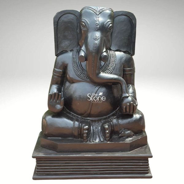 A magnificent 3.5-feet black stone Dwi-Bhuja Ganesha statue is presented from a frontal slightly elevated perspective against a plain light background. This statue depicts Lord Ganesha in a traditional serene seated posture cross-legged emphasizing his two-armed form. The entire sculpture is carved from a rich dark black stone which gives it a profound and traditional presence with a smooth polished finish that reflects light subtly. Ganesha's large expressive elephant head faces forward with his trunk curving gently towards his left. A decorative tilak or auspicious symbol is carved on his forehead and his large ears are prominently displayed. He wears a simple yet elegant crown. Unlike four-armed depictions, this Dwi-Bhuja form shows his left hand resting on his lap possibly holding an object like a modak or rosary while his right hand is raised in a gesture of blessing or teaching. He is adorned with traditional jewelry including a necklace armlets, and a sacred thread draped across his plump belly which symbolizes prosperity. His lower body is covered by finely sculpted garments with horizontal folds hinting at a dhoti. The statue rests on a solid, multi-tiered rectangular base which appears to resemble stacked books or scriptures subtly referencing Ganesha's role as the lord of wisdom and learning. This detailed image powerfully conveys the spiritual majesty and artistic craftsmanship of the black stone Dwi-Bhuja Ganesha statue, making it highly relevant to an article discussing this specific form of Ganesha.