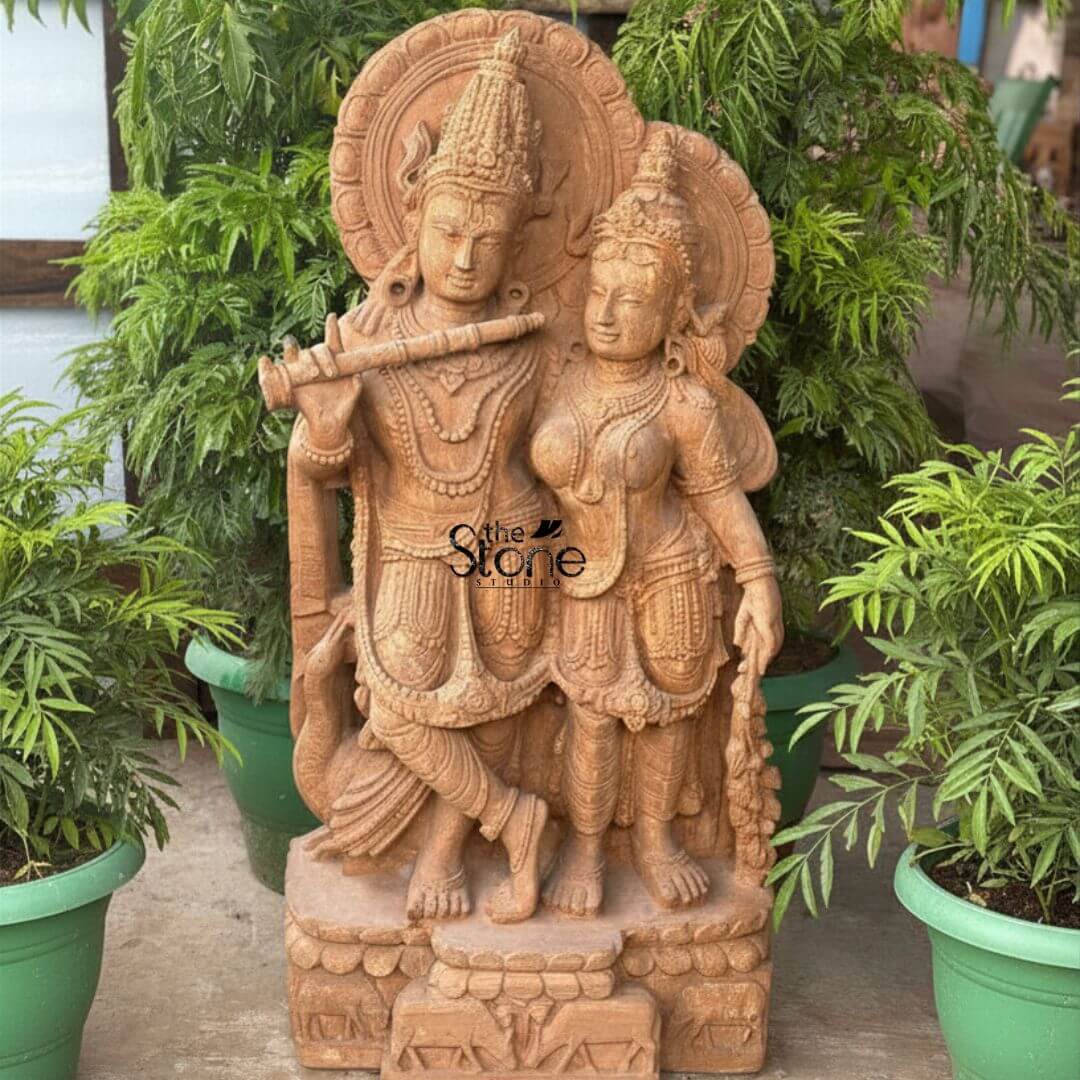 Radha Krishna Sandstone Garden Statue 3ft 3 An exquisitely hand-carved Radha Krishna sandstone sculpture masterfully crafted from premium natural brown sandstone serving as an elegant spiritual centerpiece in a lush garden setting. The sculpture captures the divine couple Lord Krishna and Goddess Radha in a traditional standing posture that radiates serenity and grace. On the left Lord Krishna is depicted in his iconic Tribhanga posture with his legs crossed delicately holding a stone flute to his lips. Radha stands elegantly to his right her gentle expression reflecting divine love. Both figures are adorned with towering tiered crowns intricate layered jewelry and circular Prabhavalis (ornate haloes) featuring delicate floral relief work. At their feet, a small detailed cow rests on a rectangular pedestal embellished with relief carvings of cattle and lotus motifs. The statue is artfully positioned in an outdoor landscape flanked by vibrant green plants in large pots set against a backdrop of dense tropical foliage. The warm earthy reddish-brown tones and rustic grainy texture of the natural sandstone give the piece a timeless antique temple aesthetic. This elegant sandstone Radha Krishna idol perfectly illustrates how traditional Indian stone art can be integrated into modern garden decor to create a peaceful sanctuary for meditation and devotion.
