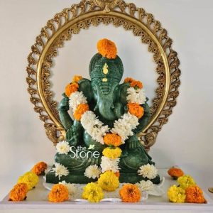A beautiful 15-inch Ganesha statue carved from green stone, is prominently displayed in a serene indoor setting suitable for an article about Ganesha statues in Pune. Lord Ganesha is seated gracefully adorned with several fresh garlands of white chrysanthemums and vibrant orange marigolds draped around his neck and chest. His trunk is gently curved to his left, and he holds traditional attributes in his hands though they are partially obscured by the garlands. Behind the green stone Ganesha, a striking, ornate golden halo provides a magnificent backdrop featuring intricate traditional patterns that enhance the deity's divine aura. The statue is placed on a pristine white surface which is also adorned with numerous loose orange and yellow marigold petals and white chrysanthemum flowers scattered around Ganesha's base creating a festive and devotional atmosphere. The subtle green hue of the stone gives the statue a unique and calming aesthetic contrasting beautifully with the golden halo and colorful flowers. The detailed carving of Ganesha's features, crown, and ornaments is evident, reflecting fine craftsmanship. This setup perfectly conveys the reverence and celebratory spirit associated with Ganesha, making it an ideal visual for an article on Ganesha statues.