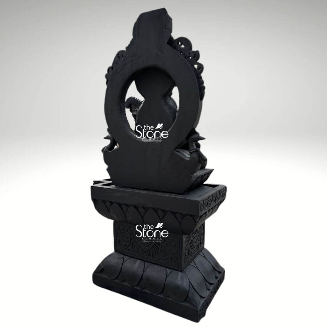 7ft Black Stone Saraswati Statue 10 A rear three-quarter view of a grand 7-foot tall Saraswati statue masterfully hand-carved from natural black stone. This perspective highlights the monumental scale and exceptional 360-degree craftsmanship of the sculpture, showcasing the solid, architectural depth of the piece. The black stone Saraswati idol is depicted seated behind a majestic circular stone arch which frames her silhouette and adds a sense of divine power and permanence. The goddess is shown atop a massive, multi-tiered pedestal that is a work of art in itself. The lowest tier features high-relief lotus-petal carvings while the central block of the base is embellished with intricate traditional floral and scrollwork relief patterns. The refined matte charcoal finish of the stone highlights the heritage of Indian stone masonry creating a timeless temple-inspired aesthetic. Standing at an impressive 7 feet, this large-scale spiritual sculpture is designed as a profound centerpiece for a grand foyer, educational institution, or temple sanctuary, inviting an atmosphere of wisdom, creativity, and divine stability into any large environment.