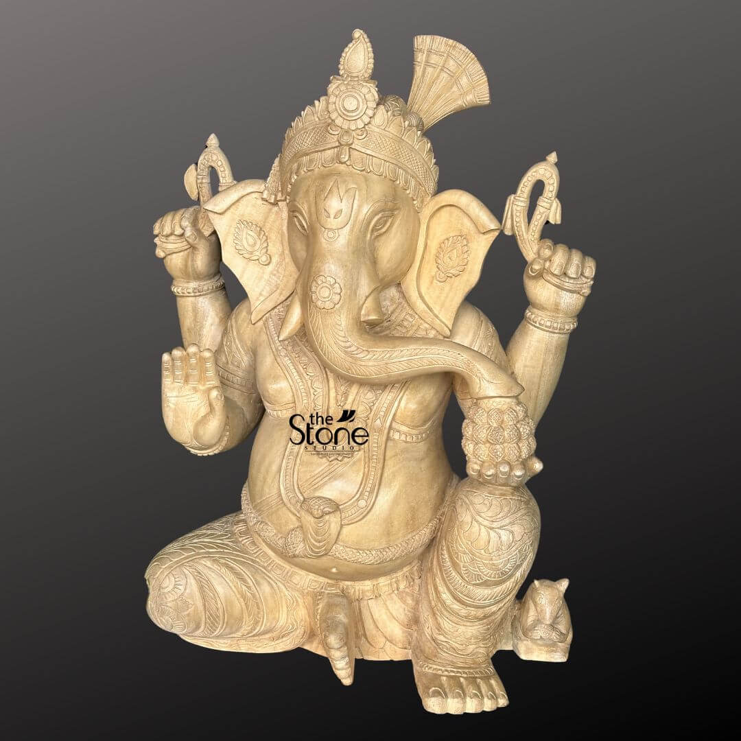 Wooden Ganesha Statue Online 2ft 4 A masterfully hand-carved 2-foot tall wooden Ganesha statue sculpted from natural light-toned wood with a smooth polished finish. This Ganpati idol is depicted in a stable seated posture showcasing a serene meditative expression with large fan-like ears and a gracefully curved trunk reaching for a bowl of modaks held in his lower left hand. Lord Ganesha’s four arms carry traditional Vedic symbols, including a goad and a noose while his lower right hand is raised in the Abhaya Mudra the universal gesture of protection and blessing. The sculpture is rich in detail featuring an intricately carved tiered crown layered necklaces armbands, and a traditional dhoti with rhythmic fabric folds. At the base of the statue, a small figure of his loyal vehicle the mouse is delicately carved near his foot. This 24-inch wooden sculpture perfectly represents traditional Indian artisan techniques making it an ideal spiritual centerpiece for a home mandir foyer, or living room to invite peace and prosperity.