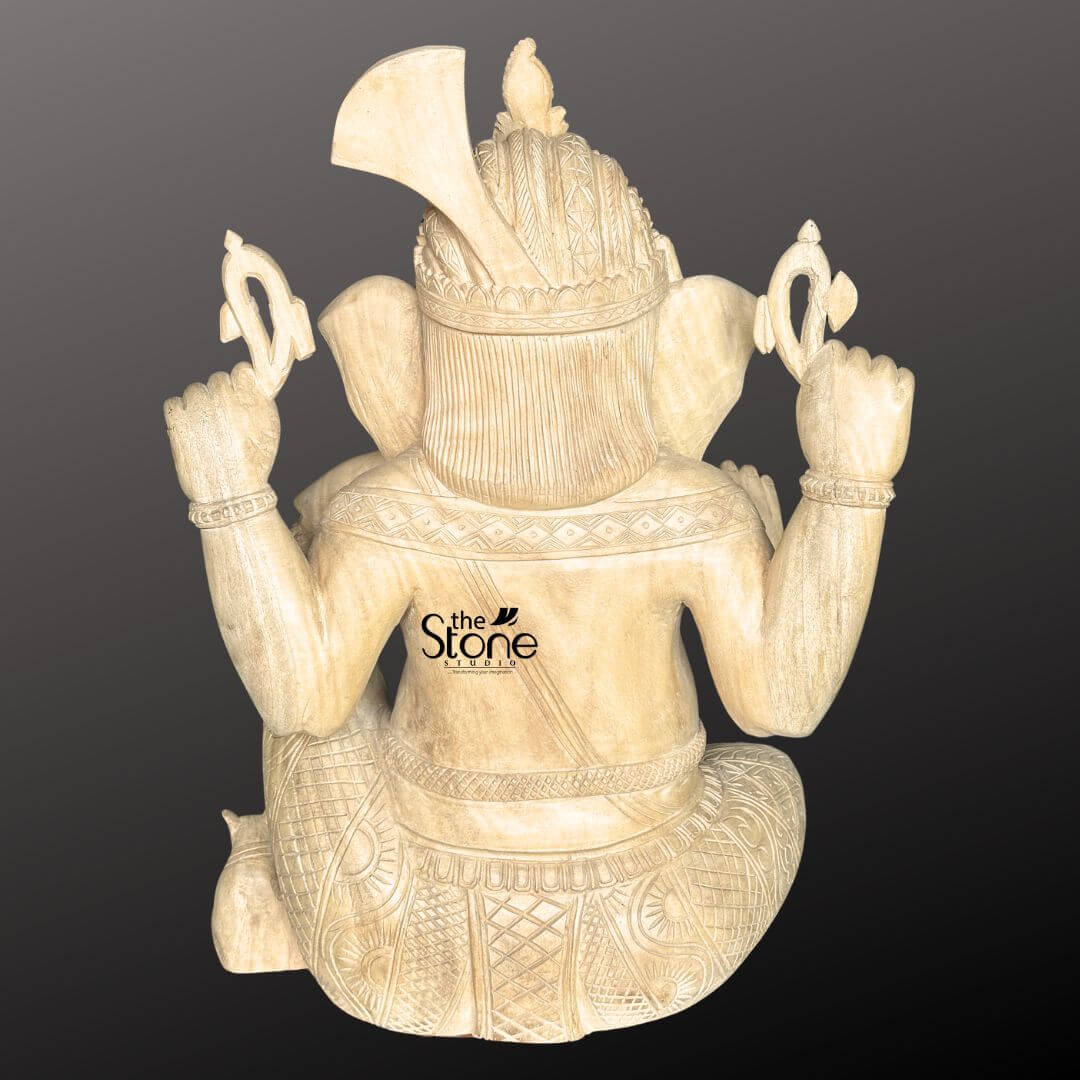 Wooden Ganesha Statue Online 2ft 5 A rear-view slightly elevated shot captures a beautifully hand-carved 2-foot wooden Ganpati idol set against a plain dark background. The statue is crafted from light-colored wood showcasing its natural grain and warm inviting tones. Lord Ganesha is depicted in a traditional seated posture with his back to the viewer emphasizing the intricate carvings on his back and attire. Ganesha's large, fan-like ears are visible and his crown or headpiece adorned with a distinctive fan-shaped element at the back is clearly detailed. His two upper hands are raised each holding stylized traditional attributes possibly a noose and an axe with detailed patterns carved into them. His back is adorned with intricate geometric and traditional patterns that simulate rich fabric or jewelry. A sacred thread is draped across his back and his lower body is covered by finely sculpted garments with horizontal folds and textured patterns. The natural wood finish adds to its rustic and handcrafted charm. This detailed image vividly conveys the artistic craftsmanship and the intricate rear details of the wooden Ganesha statue making it highly relevant to an article discussing hand-carved Ganpati wooden idols.