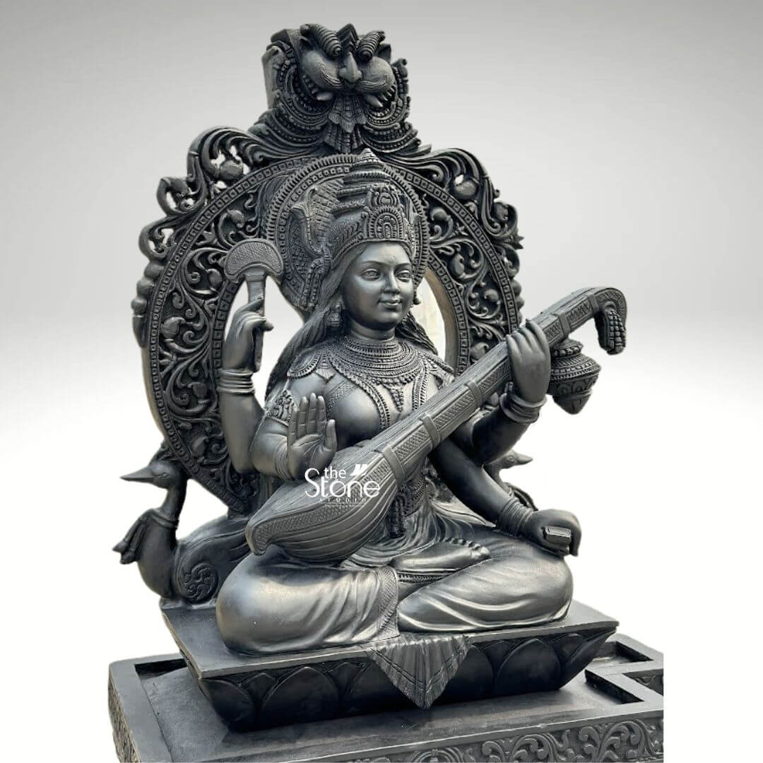7ft Black Stone Saraswati Statue 7 A magnificent 7-foot handcarved Veena Saraswati Sculpture crafted from black stone, is presented from a frontal slightly elevated perspective against a plain light background. The statue depicts Goddess Saraswati, the deity of knowledge, music, and arts, seated gracefully in a traditional cross-legged posture. She has four arms her two primary hands delicately play the elaborately carved veena held across her lap. Her upper right hand holds a rosary, and her upper left hand holds a manuscript symbolizing learning and contemplation. The entire sculpture is meticulously carved from dark black stone which imparts a profound and timeless presence. Saraswati is richly adorned with an elaborate crown, intricate necklaces, armlets, earrings, and bangles, all rendered with exceptional detail. Her garments are sculpted with delicate folds and traditional patterns. Behind her head rises a grand, arching prabhavali featuring exquisite floral, scrollwork, and possibly auspicious carvings, topped with a detailed motif, possibly a Kirtimukha. On either side of the base of the prabhavali, stylized hamsas her traditional vehicle, are depicted, further emphasizing her iconography. The entire sculpture rests on a multi-tiered rectangular base, also intricately carved with patterns. The smooth yet detailed finish of the black stone highlights the fine artistry and the serene expression of the goddess. This detailed image powerfully conveys the spiritual majesty and artistic craftsmanship of the 7-foot black stone Veena Saraswati statue, making it highly relevant to an article discussing large-scale, handcarved sculptures.