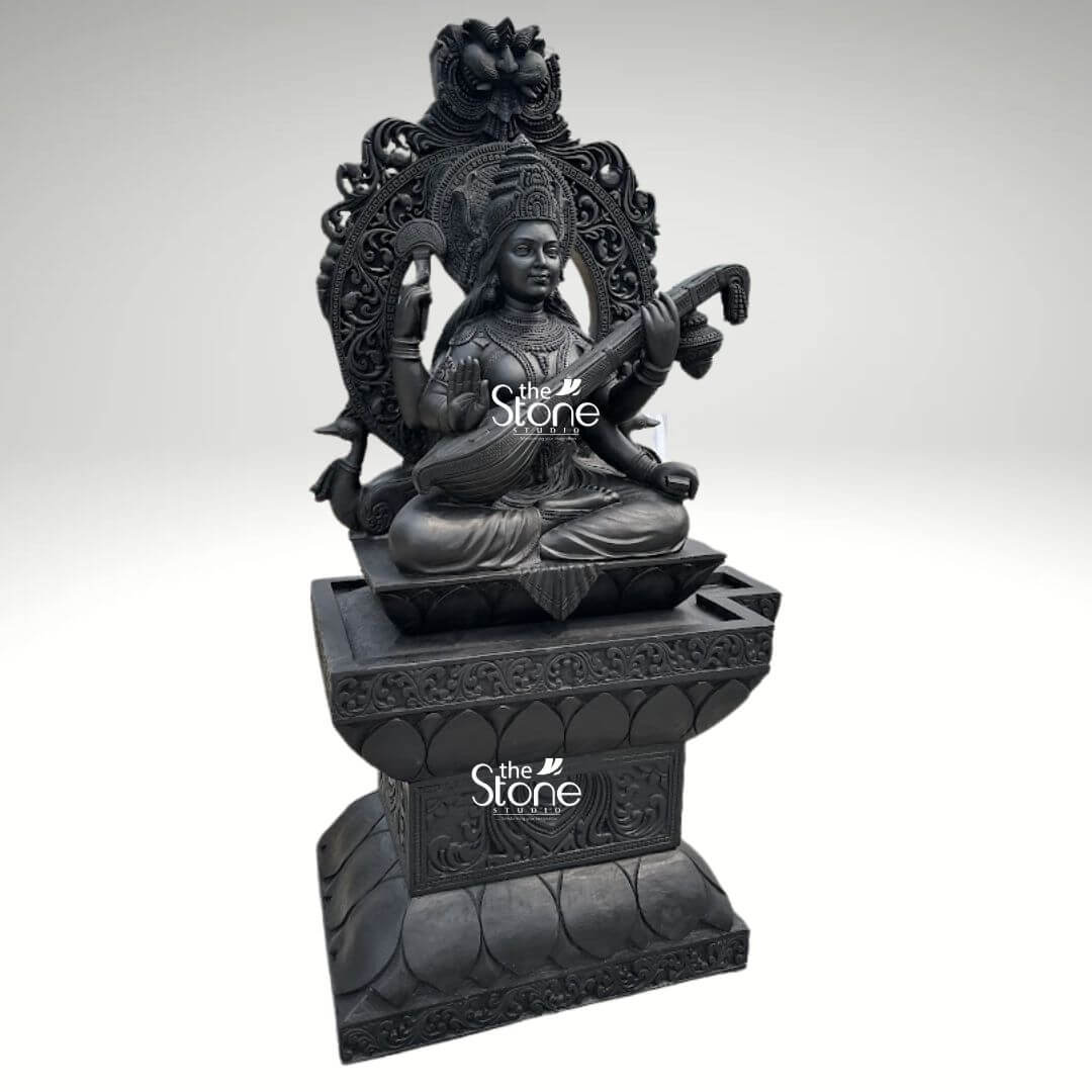 7ft Black Stone Saraswati Statue 8 A majestic 7-foot tall handcrafted Hindu Goddess Saraswati sculpture expertly carved from natural black stone with a refined matte charcoal finish. This large-scale spiritual masterpiece depicts the Goddess of knowledge, music, and the arts in a serene seated posture atop a grand, multi-tiered pedestal embellished with lotus-petal relief work. Lord Saraswati is shown with four arms, holding a sacred manuscript representing the Vedas and a prayer rosary, while her front hands gracefully play the Veena symbolizing the harmony of all creative arts. She is framed by an intricate and massive Prabhavali featuring deep relief carvings of traditional floral motifs and a protective Kirtimukha face at the peak. The craftsmanship highlights exceptional detail in her towering tiered crown layered temple jewelry, and the realistic, fluid folds of her traditional attire. This monumental black stone Saraswati statue serves as a profound spiritual and artistic centerpiece, perfect for grand temple foyers, educational institutions, or luxury home entrances, designed to invite an aura of wisdom, peace, and intellectual clarity into the environment.