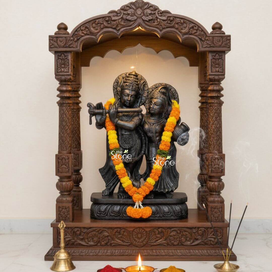 2ft Radha Krishna Black Stone Statue 3 A serene and spiritually vibrant home temple setup featuring a handcrafted 2-foot tall black stone Radha Krishna statue as the central focus. The intricately carved idol depicts Lord Krishna in a graceful standing posture playing his iconic flute, while Goddess Radha stands elegantly by his side. Both deities are adorned with a lush garland of vibrant yellow and orange marigolds highlighting the deep charcoal-black finish of the natural stone. The statue is housed within a grand dark wood mandir featuring ornate pillars and traditional floral relief carvings on the archway. The devotional setting is further enhanced by ritual items in the foreground including a polished brass puja bell a lit oil lamp (diya) casting a warm glow and burning incense sticks with delicate wisps of smoke rising into the air. Placed upon a smooth white marble floor this 2ft black stone Radha Krishna sculpture perfectly illustrates the fusion of ancient stone craftsmanship and modern spiritual decor. This setup serves as an ideal inspiration for creating a peaceful sanctuary for daily prayer and meditation inviting an aura of divine love and prosperity into a contemporary home mandir.