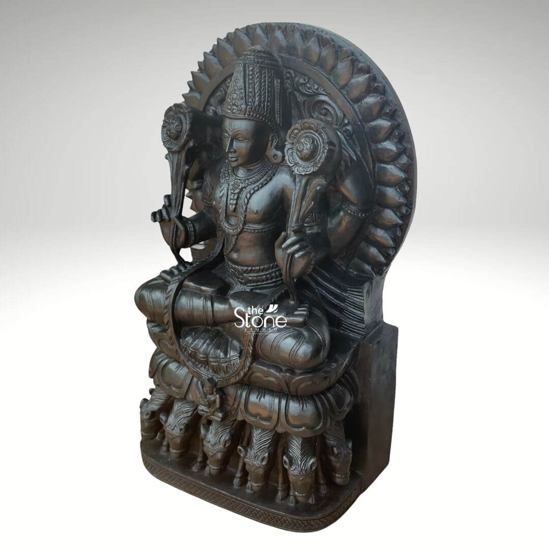Surya Dev Black Stone Statue 30" 5 An exquisitely handcrafted 2.5-foot tall Surya Dev murti masterfully hand-carved from natural black stone serves as a majestic spiritual centerpiece. Shown from a three-quarter side angle to highlight its depth and volume this black stone Surya Dev statue depicts the Sun God in a traditional seated posture, radiating an aura of divine energy and cosmic discipline. The deity is portrayed with two arms, each holding a long-stemmed, blooming lotus flower which symbolizes purity and the awakening of consciousness. Lord Surya is adorned with a towering, tiered mukut and intricate temple jewelry, including layered necklaces and armlets that showcase the exceptional craftsmanship of traditional Indian stone masonry. Framing the head and upper body is a grand, circular Prabhavali embellished with rhythmic petal-like relief patterns that represent the life-giving rays of the sun. The entire 30-inch stone sculpture rests upon a multi-tiered pedestal. A defining iconographic feature is visible at the base: the seven horses of his celestial chariot are meticulously carved in high relief, representing the seven days of the week and the seven colors of light. The refined matte charcoal finish of the stone highlights the heritage and permanence of this handcrafted stone idol. This durable and weather-resistant Surya Dev statue is an ideal addition to a home mandir, grand foyer, or office space, designed to invite vitality, success, and positive energy into the surroundings.