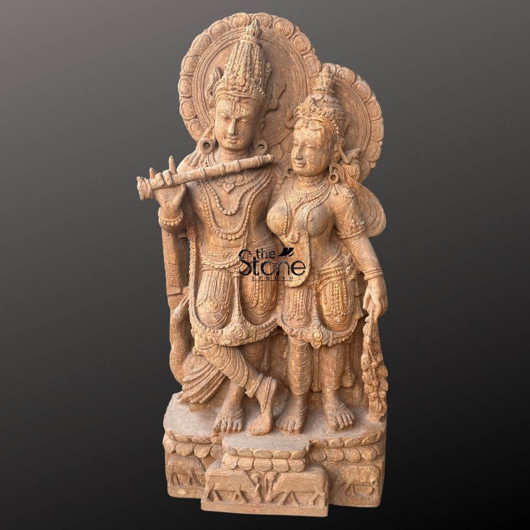Radha Krishna Sandstone Garden Statue 3ft 4 An exquisitely detailed handmade Radha Krishna sandstone statue masterfully hand-carved from natural brown stone with a rich grainy texture. The sculpture depicts the divine couple in a traditional standing pose Lord Krishna is seen on the left in his iconic Tribhanga posture with legs crossed gracefully holding a flute to his lips. To his right Goddess Radha stands with a gentle, devoted expression looking toward him. Both figures are adorned with towering tiered crowns, layered necklaces, armlets, and circular Prabhavalis featuring rhythmic sunburst and floral patterns. The entire idol rests on a sturdy rectangular pedestal embellished with relief carvings of cows and lotus motifs symbolizing purity and the divine cowherd's life. This sandstone Radha Krishna idol serves as a timeless spiritual centerpiece for a home mandir, luxury garden, or meditation sanctuary, inviting an aura of peace and divine love into the surrounding environment.