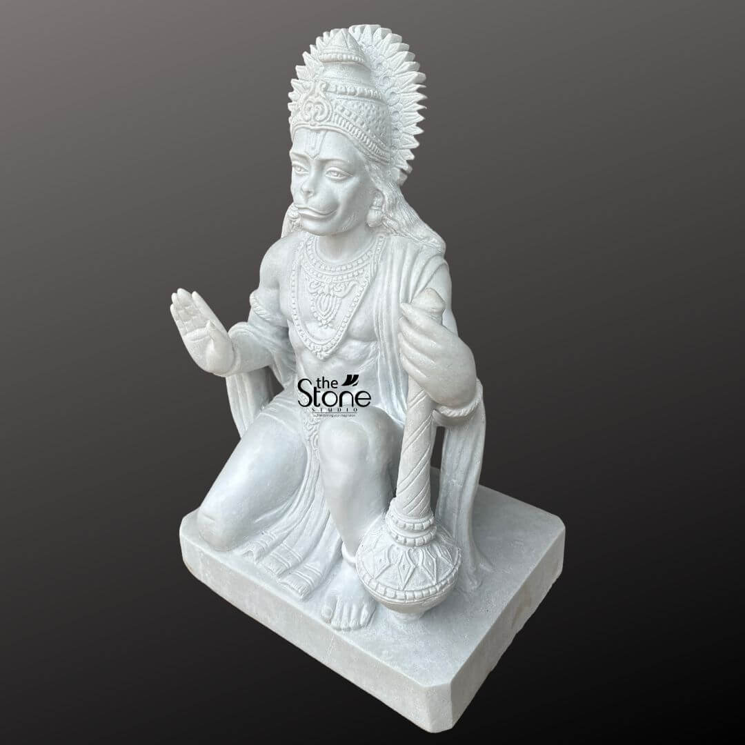 Hanuman Marble Statue for Home Temple 3ft 5 An exquisitely hand-carved 3-foot tall Lord Hanuman Ji marble murti depicted in a heroic kneeling posture serves as a profound spiritual centerpiece for a home temple or sanctuary. Sculpted from premium pure white marble with a smooth, polished finish, this Hanuman Ji marble statue radiates a sense of divine strength loyalty, and peace. The deity is shown with his right hand raised in the Abhaya Mudra the universal gesture of fearlessness and blessing, offering divine protection to all who behold it. In his left hand he firmly holds his signature weapon, the Gada positioned vertically on the floor. Masterful craftsmanship is highlighted throughout the piece, including a towering sunburst-style mukut detailed layered necklaces, armlets, and the realistic, fluid folds of his traditional dhoti. The serene facial expression and a prominent red tilak on his forehead convey deep spiritual wisdom. The entire sculpture sits firmly upon a solid, square marble pedestal, embodying stability and permanence. This large-scale 36-inch white marble Hanuman statue perfectly blends classical Vedic iconography with expert stone artistry, creating an auspicious and tranquil atmosphere ideal for daily devotion, meditation, and mindfulness.
