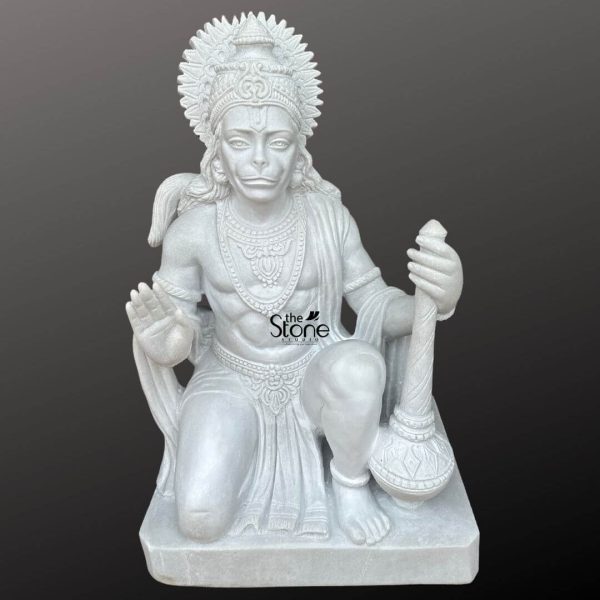 A commanding 3-foot marble Hanuman statue perfect for a home temple is presented from a frontal slightly elevated perspective against a plain dark background. The statue is sculpted entirely from pristine white marble which highlights every fine detail with remarkable clarity and luminosity. Lord Hanuman is depicted in a dynamic kneeling posture poised for action yet radiating a calm and devoted expression. His right hand is raised in a gesture of blessing or fearlessness offering protection while his left hand rests on his knee firmly grasping his gada, a symbol of immense strength discipline and devotion. Hanuman's face is rendered with distinct simian features including expressive eyes and a slightly pointed snout yet conveying profound wisdom loyalty and unwavering devotion. He is richly adorned with an elaborate crown or headpiece featuring a sun-like halo with radiating rays intricate necklaces armlets anklets and a sacred thread draped across his muscular chest. His powerful physique is subtly sculpted covered by finely draped garments that show detailed folds and patterns. The entire statue rests on a simple rectangular marble base. The smooth lustrous finish of the white marble emphasizes the exquisite artistry and the powerful benevolent and protective presence of Lord Hanuman. This detailed image vividly conveys the spiritual majesty and artistic craftsmanship of the marble Hanuman statue making it highly relevant to an article discussing such revered idols for home temple use.