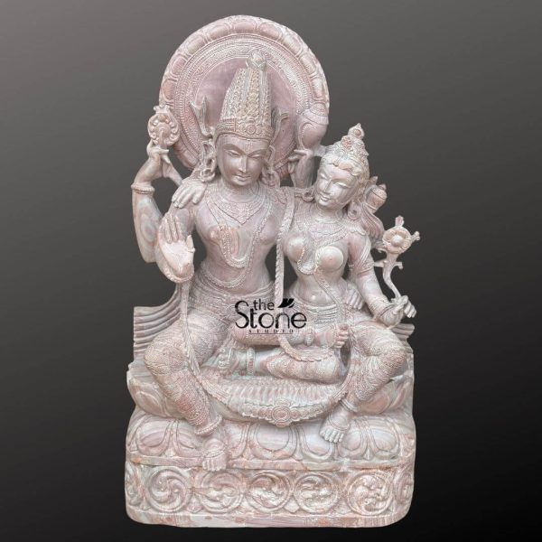 An exquisitely hand-carved 3-foot tall Lakshmi Narayana Perumal statue masterfully sculpted from natural pinkstone with a rich grainy texture. This detailed sculpture captures the divine union of Lord Vishnu and Goddess Lakshmi in a serene and harmonious seated posture, radiating an aura of peace and spiritual grace. Lord Vishnu is portrayed on the left with four arms, holding his sacred symbols—the Sudarshana Chakra and the Panchajanya Shankha —in his upper hands. His lower right hand is raised in the Abhaya Mudra the universal gesture of protection and blessing, while Goddess Lakshmi sits gracefully by his side, her gentle expression reflecting abundance and devotion. Both figures are adorned with towering, multi-tiered crowns intricate layered necklaces and armlets that showcase the exceptional craftsmanship of traditional Indian stone masonry. The sculpture features highly detailed relief work including a grand circular Prabhavali with rhythmic floral and sunburst patterns framing their heads. The realistic fluid fabric folds of their traditional attire add a sense of lifelike elegance to the solid stone. The entire pinkstone Laxmi Narayan murti rests on a wide multi-tiered pedestal embellished with lotus petal relief carvings and scrolling vine motifs at the base. The warm, earthy rose tones and rustic finish of the natural stone provide a timeless antique temple aesthetic making this large-scale idol ideal for a home mandir, office foyer, or meditation sanctuary to invite divine harmony, prosperity, and protection into the environment.