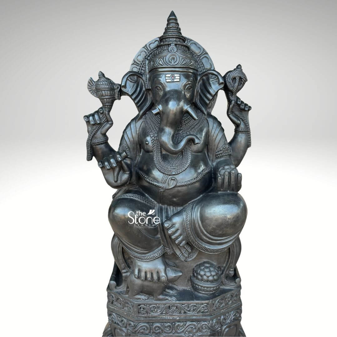 3.5ft Ganesha Garden Statue 5 This image presents a magnificent 3.5-foot black stone Ganesha statue a truly large and commanding piece displayed against a plain light background that highlights its sculptural details. The deity is seated in a traditional relaxed posture exuding an aura of peace and wisdom. Ganesha's distinctive elephant head is carved with remarkable precision, featuring broad fanned ears deep-set, serene eyes and a trunk gracefully curled towards his left symbolizing prosperity and the ability to overcome obstacles. Adorning his forehead is a prominent Tilak mark adding to his divine countenance. He wears an elaborate crown multiple necklaces and detailed armlets and anklets all meticulously sculpted to showcase the rich texture and depth of the black stone. The large statue is enthroned on a beautifully designed multi-layered lotus pedestal, a sacred symbol of purity and spiritual enlightenment. This pedestal, in turn rests upon a sturdy octagonal base which is intricately embellished with delicate floral and geometric carvings further enhancing the statue's aesthetic and cultural value. Ganesha's four hands each hold significant attributes: his upper right hand grasps a conch shell and his upper left an axe representing divine sound and the power to remove impediments. His lower right hand is raised in a gesture of blessing, offering grace and protection while his lower left hand cradles a bowl of modak, his favorite sweet, symbolizing the sweet fruits of devotion and knowledge. The smooth dark finish of the black stone imparts a timeless elegance and powerful presence to this Large 3.5ft Ganesha Statue. Every curve and contour, from the folds of his dhoti to the delicate details of his jewelry, is rendered with exceptional craftsmanship. This detailed alt text conveys a comprehensive visual description, emphasizing the statue's impressive size and intricate artistry, making it clear why such a piece would be a focal point in any discussion about substantial and aesthetically rich Ganesha sculptures.