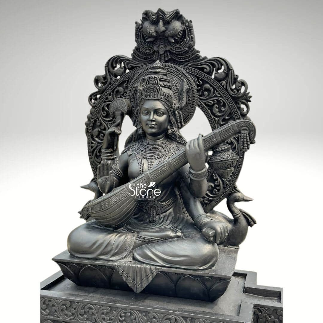 7ft Black Stone Saraswati Statue 6 A grand 7-foot black stone Saraswati statue is presented from a frontal slightly elevated perspective against a plain light background. The monumental sculpture depicts Goddess Saraswati the Hindu deity of knowledge, music, arts, and learning, seated in a serene, traditional cross-legged posture (Padmasana). She has four arms; her two primary hands delicately play the intricately carved veena held across her lap. Her upper right hand holds a rosary symbolizing meditation and spiritual knowledge, and her upper left hand holds a manuscript, representing books and learning. The entire statue is meticulously carved from dark black stone, which imparts a powerful, timeless, and traditional presence. Saraswati is richly adorned with an elaborate crown, intricate necklaces, armlets, earrings, and bangles, all rendered with exceptional detail and texture. Her garments are sculpted with delicate folds and traditional patterns, emphasizing her graceful form. Behind her head rises a magnificent, arching prabhavali featuring exquisite floral, scrollwork, and geometric carvings, topped with a detailed motif, possibly a Kirtimukha or a symbolic animal. On either side of the base of the prabhavali, stylized hamsas her traditional vehicle, are depicted, further emphasizing her iconography. The entire sculpture rests on a multi-tiered rectangular base, also intricately carved with patterns along its sides. The smooth yet detailed finish of the black stone highlights the fine artistry and the serene, benevolent expression of the goddess. This detailed image powerfully conveys the spiritual majesty and artistic craftsmanship of the large 7-foot black stone Saraswati statue, making it highly relevant to an article discussing such grand sculptures for temples or prominent spaces.