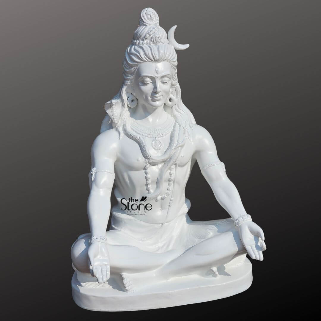 2ft Shiva Meditation Statue 6 A majestic 2-foot fiber Lord Shiva Sitting Statue perfect for home placement is presented from a slightly elevated frontal perspective against a plain dark background. The statue is entirely white symbolizing purity and transcendence. Lord Shiva is depicted in a profound meditative posture (Padmasana or Siddhasana), with his legs crossed and his hands resting open in his lap palms facing upwards likely in Dhyana Mudra (meditation gesture). His eyes are gently closed or downcast, conveying deep inner calm and spiritual introspection. Shiva is adorned with his traditional iconography a prominent crescent moon is nestled in his intricately matted hair (Jata), which flows down his shoulders. A finely sculpted cobra is draped around his neck and shoulders, and he wears multiple strands of rudraksha beads as necklaces and armlets. His third eye is subtly marked on his forehead, and large earrings dangle from his ears. The smooth, lustrous finish of the fiber material highlights every detail of his powerful yet serene form. The statue rests on a simple, oval-shaped white base, completing its elegant presentation. This detailed image effectively conveys the spiritual essence and artistic craftsmanship of the fiber Lord Shiva sitting statue, making it highly relevant to an article discussing home idols.