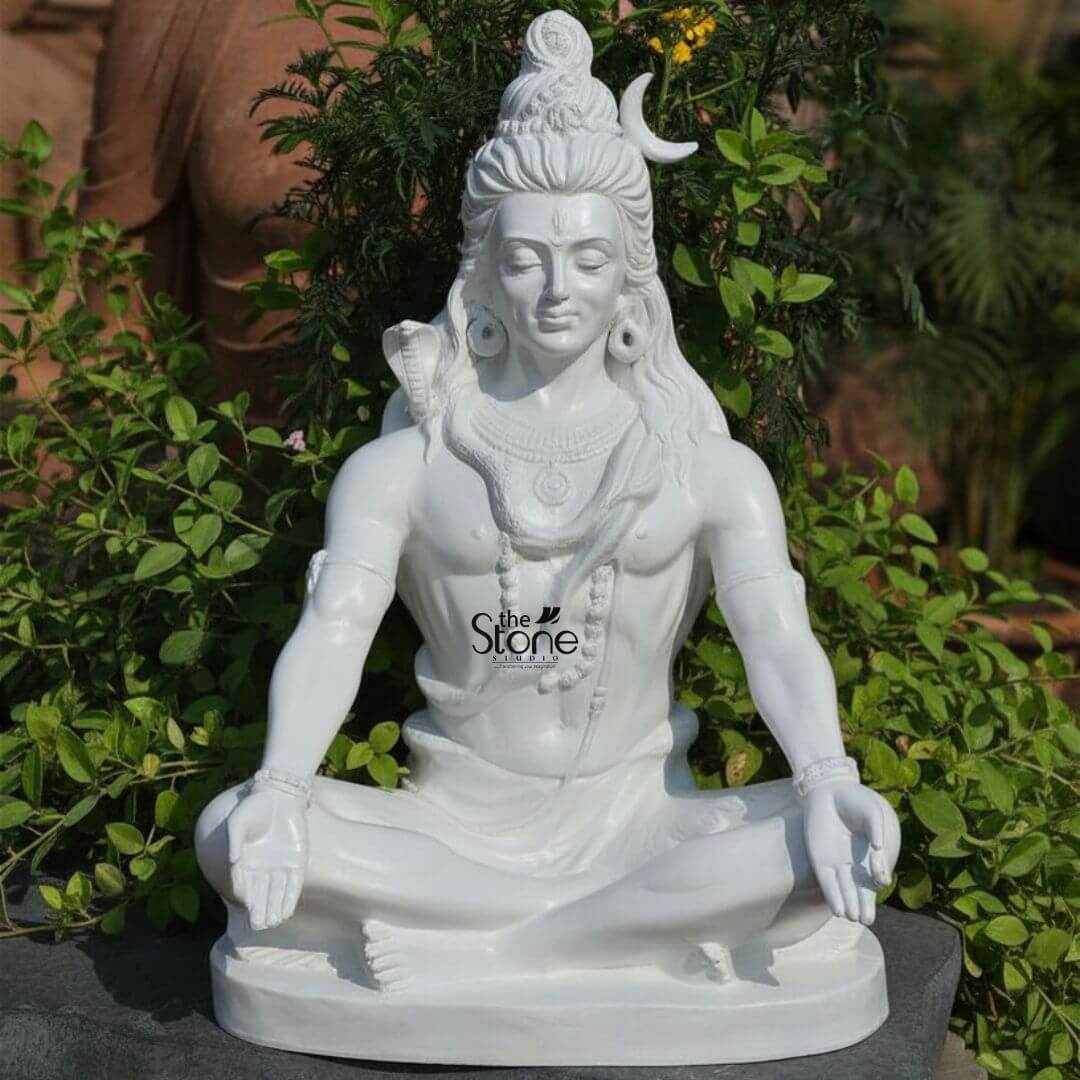 2ft Shiva Meditation Statue 3 A striking 2-foot fiber Lord Shiva statue ideal for home worship is beautifully situated in an outdoor garden setting amidst lush green foliage. The statue is entirely white symbolizing purity, peace, and divine power. Lord Shiva is depicted in a deep meditative seated posture (Padmasana), with his legs crossed and his hands resting open in his lap, palms facing upwards, in Dhyana Mudra. His eyes are gently closed or downcast, conveying profound inner calm and spiritual introspection. Shiva is richly adorned with his traditional attributes: a prominent crescent moon is nestled in his intricately matted hair (Jata), which flows gracefully down his shoulders. A finely sculpted cobra is draped around his neck and shoulders and he wears multiple strands of rudraksha beads as necklaces and armlets. His third eye is subtly marked on his forehead and large earrings adorn his ears. The smooth, lustrous finish of the fiber material highlights every detail of his powerful yet serene form. The statue rests on a simple, flat dark gray base, contrasting subtly with the white of the idol and the surrounding greenery. This detailed image effectively conveys the spiritual essence and artistic craftsmanship of the fiber Lord Shiva statue making it highly relevant to an article discussing home idols and their placement.