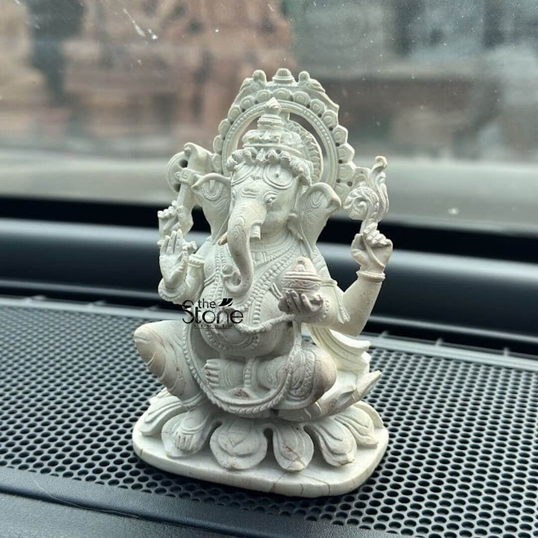 Small Ganesha Idol for Car Dashboard 4" 3 A small intricately carved white stone Ganesha statue marketed as a lucky Ganpati car statue is positioned on the textured dashboard of a vehicle. The Ganesha idol is seated in a traditional posture on a lotus base radiating an aura of protection and auspiciousness. It features four arms one upper hand holds an axe or goad another holds a noose a lower hand is raised in a blessing gesture and the remaining hand holds a bowl of modaks with the trunk curving towards it. The white stone material possibly with natural veining, highlights the fine details of the crown, jewelry, and the folds of the dhoti. A decorative, flame-like halo or prabhavali with a serrated edge frames Ganesha's head. The dashboard's textured surface and the slightly blurred view through the windshield in the background clearly indicate its placement within a car. This detailed image effectively conveys the spiritual and decorative purpose of the white stone Ganesha statue for car use aligning with the article's content about lucky car idols.
