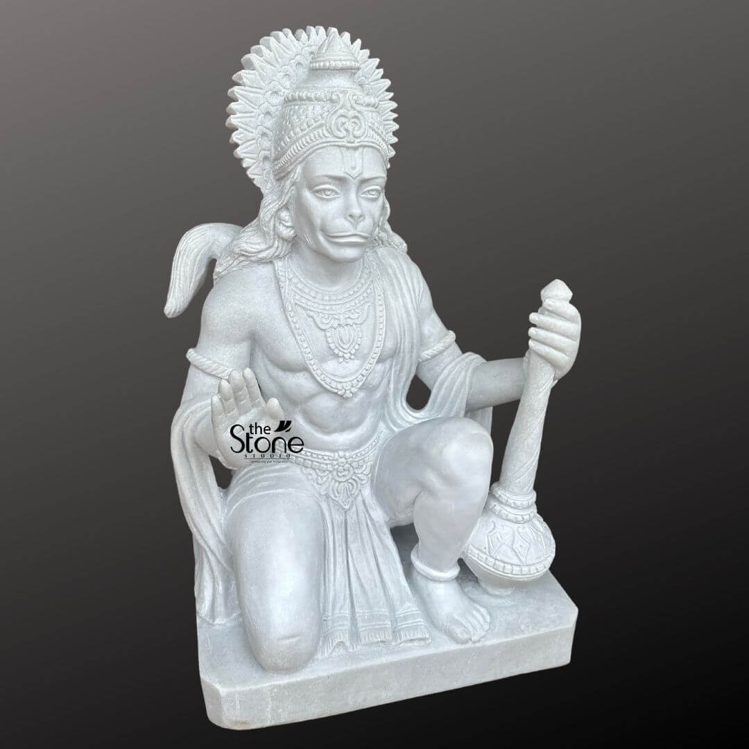 Hanuman Marble Statue for Home Temple 3ft 7 A powerful and intricately carved 3-foot marble Sankat Mochan Hanuman Statue ideal for home worship is presented from a frontal slightly elevated perspective against a plain dark background. The statue is sculpted entirely from pristine white marble which highlights every fine detail with remarkable clarity and luminosity. Lord Hanuman known as Bajrangbali or Sankat Mochan is depicted in a dynamic kneeling posture poised for action yet radiating a calm and devoted expression. His right hand is raised in a gesture of blessing or fearlessness offering protection while his left hand rests on his knee, firmly grasping his gada a symbol of immense strength, discipline, and devotion. Hanuman's face is rendered with distinct simian features including expressive eyes and a slightly pointed snout yet conveying profound wisdom, loyalty, and unwavering devotion. He is richly adorned with an elaborate crown or headpiece featuring a sun-like halo with radiating rays, intricate necklaces, armlets, anklets, and a sacred thread (yajnopavita) draped across his muscular chest. His powerful physique is subtly sculpted, covered by finely draped garments that show detailed folds and patterns. The entire statue rests on a simple, rectangular marble base. The smooth, lustrous finish of the white marble emphasizes the exquisite artistry and the powerful, benevolent, and protective presence of Lord Hanuman. This detailed image vividly conveys the spiritual majesty and artistic craftsmanship of the marble Sankat Mochan Hanuman statue making it highly relevant to an article discussing such revered idols for home use.