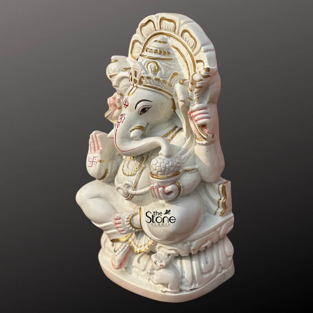 Marble Stone Ganesha Statue 15" 2 A vibrant 12-inch Ganesha statue crafted from white marble is showcased from a slightly elevated side angle against a dark gradient background perfect for an article about Marble Stone Ganapathi Statue. Lord Ganesha is seated gracefully on a lotus-like marble base which features delicate gold accents. His trunk is gently curved to his left. In his hands Ganesha holds traditional attributes: an axe in his upper right a noose in his upper left, and a modak in his lower left. His lower right hand is raised in a gesture of blessing adorned with a red swastika symbol. Red markings including an Om symbol on his forehead and detailing around his eyes and trunk add striking contrast to the pristine white marble. Ganesha is richly adorned with golden-painted jewelry including a crown necklaces armlets, and anklets enhancing his divine appearance. Behind him a beautifully carved marble prabhavali with an openwork design is highlighted with gold paint creating a radiant backdrop. At his feet his tiny mouse vahana is sculpted looking up at him. The combination of smooth white marble vibrant red and gold accents creates an auspicious and visually appealing representation of Lord Ganapathi.