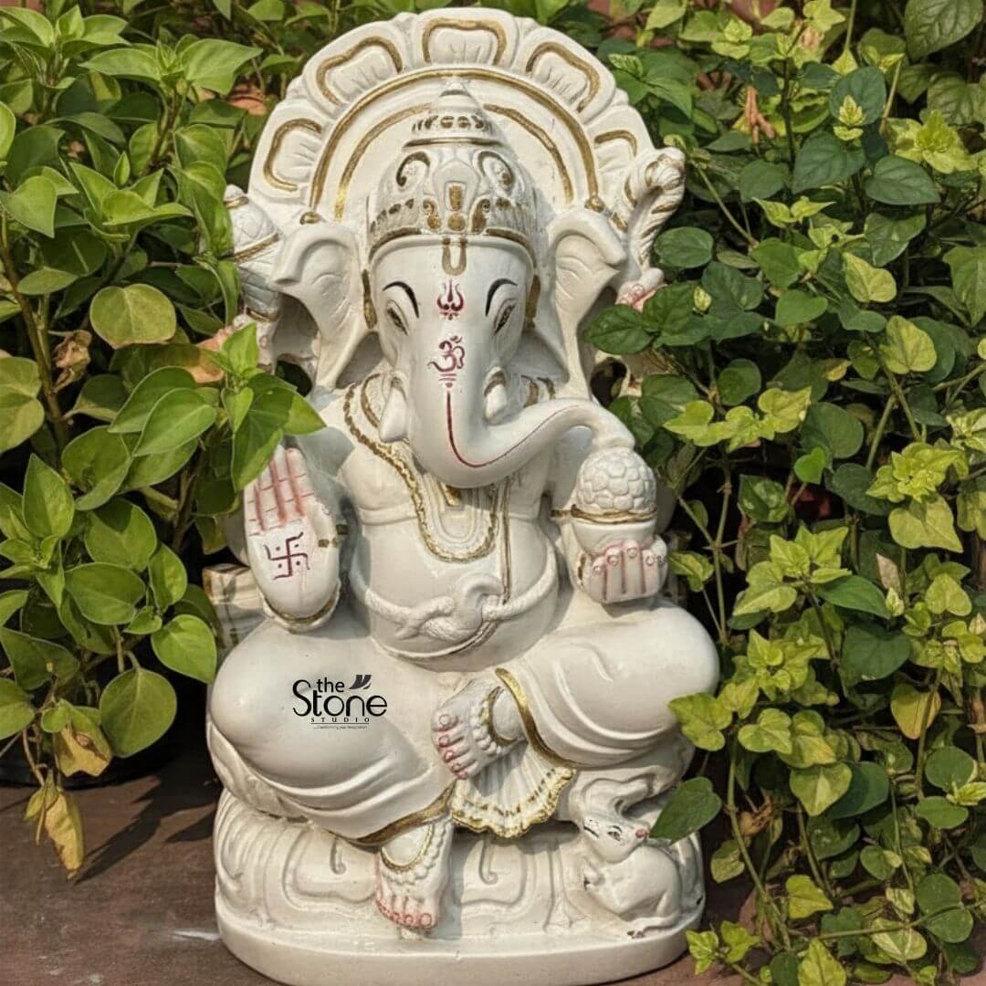 Marble Stone Ganesha Statue 15" 3 A radiant 12-inch Ganesha statue meticulously carved from white marble, is set outdoors amidst lush green foliage creating a serene environment perfect for a Marble Stone Ganesha Idol for home. Lord Ganesha is depicted seated gracefully on a lotus-like marble base which features subtle golden accents. His trunk is gently curved to his left and his four hands hold traditional attributes. In his upper right hand he holds an axe and in his upper left a noose. His lower right hand is raised in a gesture of blessing adorned with a vibrant red swastika symbol. His lower left hand holds a modak symbolizing blessings and prosperity. A prominent red Om symbol is painted on his forehead and red markings also highlight his trunk and eyes adding a beautiful contrast to the white marble. Ganesha is richly adorned with golden-painted jewelry including an elaborate crown, necklaces, armlets, and anklets, enhancing his divine appearance. Behind him, a beautifully carved marble prabhavali with a delicate openwork design is highlighted with gold paint forming a radiant backdrop. At his feet his tiny mouse vahana is sculpted, looking up at him. The surrounding dense green leaves provide a natural organic frame for the pure white and golden deity making it an ideal piece for a home altar or garden.