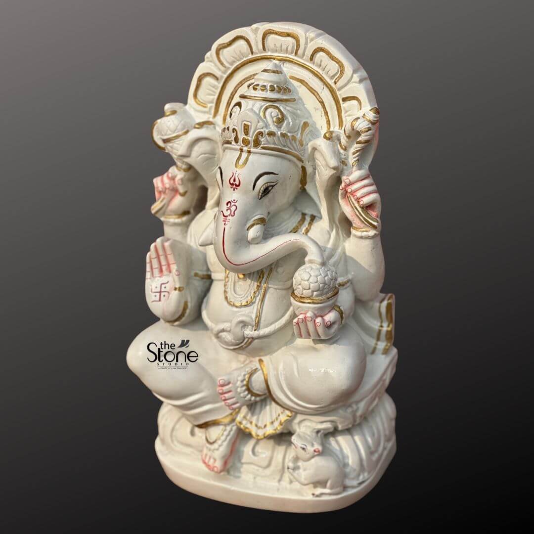Marble Stone Ganesha Statue 15" 5 A pristine 12-inch Ganesha statue meticulously carved from white marble is showcased from a slightly elevated side angle against a dark gradient background, perfect for an article about Marble Stone Ganesha Idol. Lord Ganesha is seated gracefully on a lotus-like marble base which features delicate gold accents. His trunk is gently curved to his left. In his hands Ganesha holds traditional attributes: an axe in his upper right, a noose in his upper left, and a modak in his lower left. His lower right hand is raised in a gesture of blessing adorned with a vibrant red swastika symbol. Red markings including an Om symbol on his forehead and detailing around his eyes and trunk, add striking contrast to the pristine white marble. Pink accents are also visible on his ears and fingertips. Ganesha is richly adorned with golden-painted jewelry including an elaborate crown, necklaces, armlets, and anklets enhancing his divine appearance. Behind him, a beautifully carved marble prabhavali with an openwork design is highlighted with gold paint, creating a radiant backdrop. At his feet, his tiny mouse vahana is sculpted, looking up at him. The combination of smooth white marble, vibrant red, pink, and gold accents creates an auspicious and visually appealing representation of Lord Ganesha.