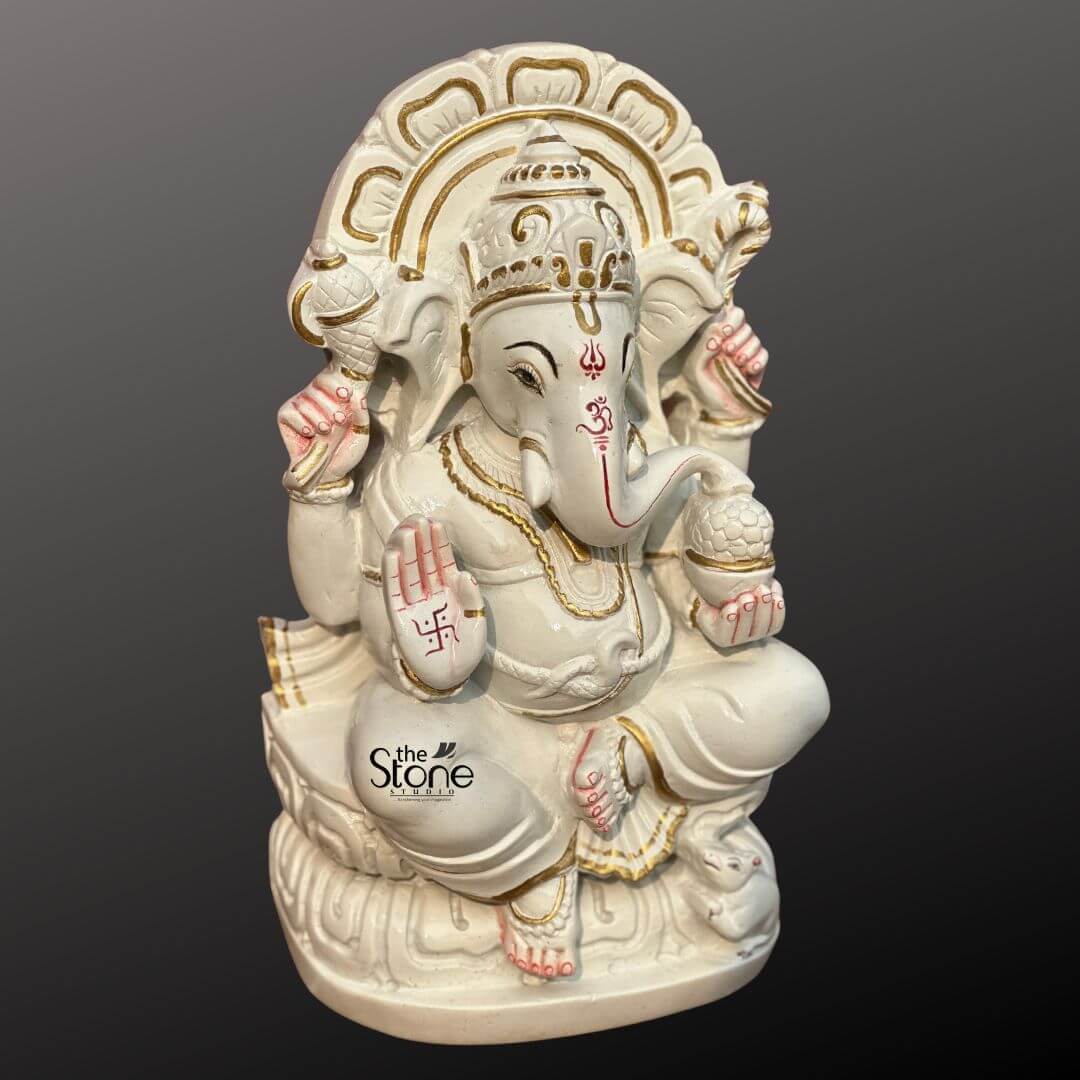 Marble Stone Ganesha Statue 15" 4 A beautiful 12-inch Ganesha statue meticulously sculpted from white marble is showcased from a slightly elevated perspective against a dark, gradient background fitting for an article on Marble Stone Ganesha Sculpture. Lord Ganesha is seated gracefully on a lotus-like marble base which is subtly accented with gold paint. His trunk is gently curved to his left and his four hands hold traditional attributes. In his upper right hand he holds an axe, and in his upper left a noose. His lower right hand is raised in a gesture of blessing featuring a vibrant red swastika symbol. His lower left hand holds a modak symbolizing blessings and spiritual rewards. A prominent red Om symbol is painted on his forehead and red markings also highlight his trunk eyes, and fingertips, adding striking contrast to the pristine white marble. Soft pink accents are also visible on his ears. Ganesha is richly adorned with golden-painted jewelry including an elaborate crown, necklaces, armlets, and anklets, enhancing his divine appearance. Behind him, a beautifully carved marble prabhavali with a delicate openwork design is highlighted with gold paint, forming a radiant backdrop. At his feet, his tiny mouse vahana is sculpted, looking up at him. The combination of smooth white marble with vibrant red, pink, and gold accents creates an auspicious and artistically appealing representation of Lord Ganesha.