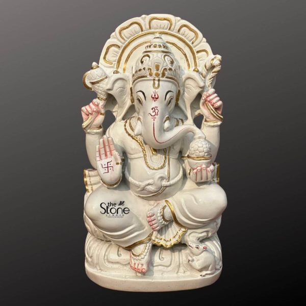 A pristine 12-inch Ganesha statue crafted from white marble is presented against a dark gradient background ideal for an article about Marble Stone Ganesha Statue. Lord Ganesha is depicted seated elegantly on a lotus-like base which is also carved from marble and features subtle gold accents. His trunk is gently curved to his left and his four hands hold traditional attributes. In his upper right hand he holds an axe and in his upper left a noose. His lower right hand is raised in a gesture of blessing featuring a red swastika symbol while his lower left hand holds a modak symbolizing rewards for spiritual seeking. A red Om symbol is painted on his forehead and red markings also highlight his trunk and eyes adding vibrant contrast to the white marble. Ganesha is adorned with golden-painted jewelry including a crown necklaces, armlets and anklets, enhancing his divine appearance. Behind him a beautifully carved marble prabhavali with a delicate openwork design is highlighted with gold paint forming a radiant backdrop. At his feet his tiny mouse vahana is sculpted looking up at him. The smooth white marble with touches of gold and red creates a classic and auspicious representation of Lord Ganesha.