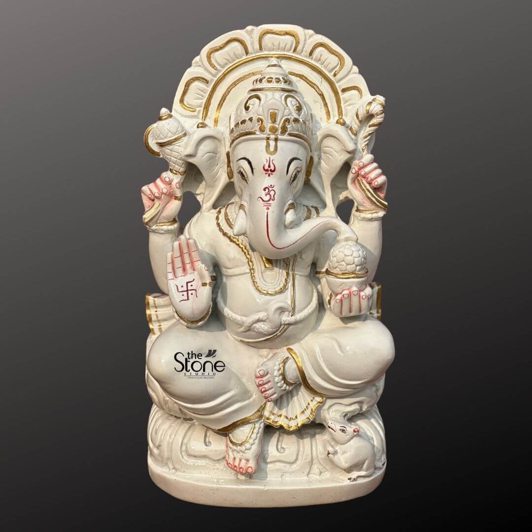 Marble Stone Ganesha Statue 15" 1 A pristine 12-inch Ganesha statue crafted from white marble is presented against a dark gradient background ideal for an article about Marble Stone Ganesha Statue. Lord Ganesha is depicted seated elegantly on a lotus-like base which is also carved from marble and features subtle gold accents. His trunk is gently curved to his left and his four hands hold traditional attributes. In his upper right hand he holds an axe and in his upper left a noose. His lower right hand is raised in a gesture of blessing featuring a red swastika symbol while his lower left hand holds a modak symbolizing rewards for spiritual seeking. A red Om symbol is painted on his forehead and red markings also highlight his trunk and eyes adding vibrant contrast to the white marble. Ganesha is adorned with golden-painted jewelry including a crown necklaces, armlets and anklets, enhancing his divine appearance. Behind him a beautifully carved marble prabhavali with a delicate openwork design is highlighted with gold paint forming a radiant backdrop. At his feet his tiny mouse vahana is sculpted looking up at him. The smooth white marble with touches of gold and red creates a classic and auspicious representation of Lord Ganesha.