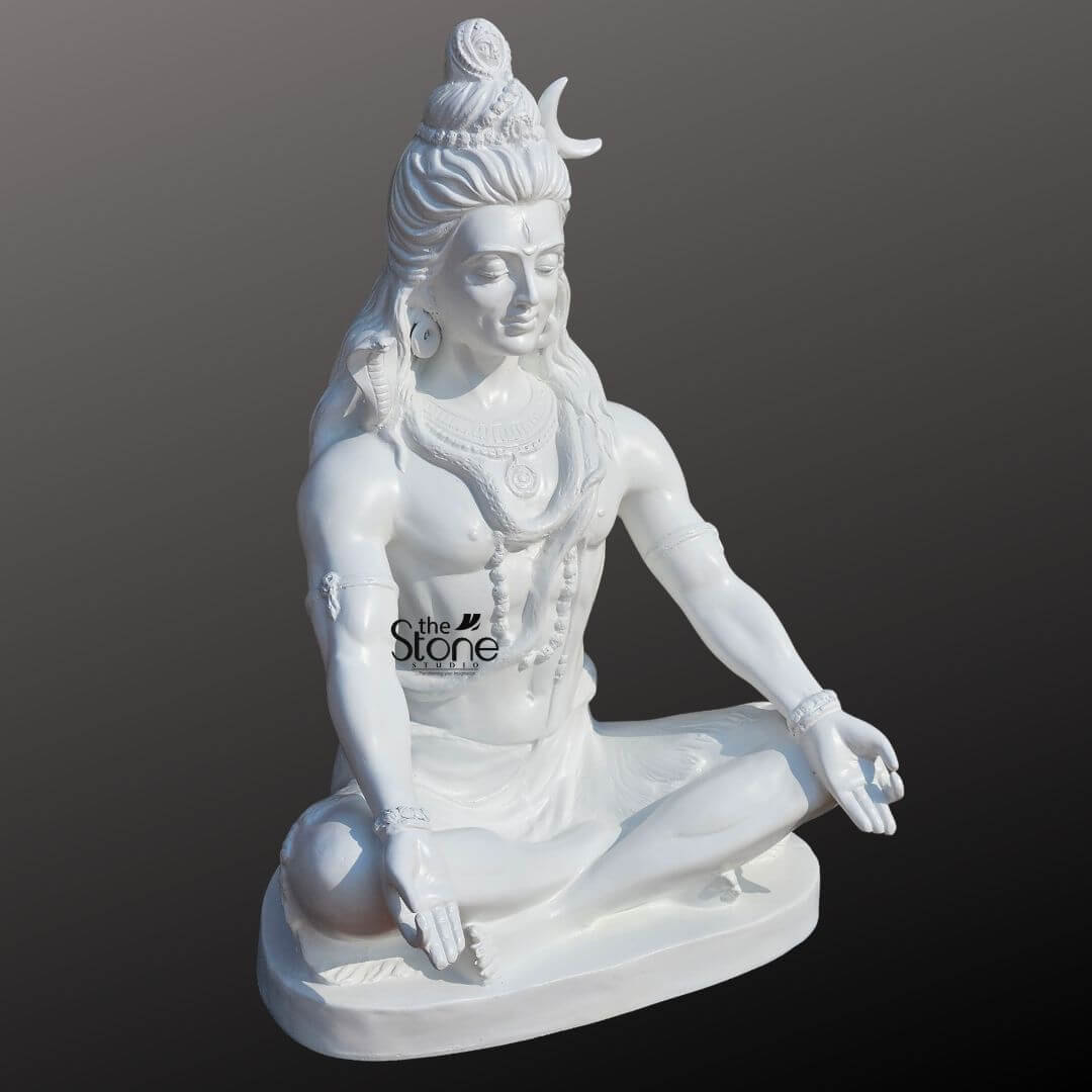 2ft Shiva Meditation Statue 8 An exquisitely hand-carved 2-foot tall meditative Shiva statue masterfully designed for spiritual home decor. Shown from a serene three-quarter side angle the sculpture captures Lord Shiva in a state of deep contemplation seated cross-legged in the traditional Padmasana (lotus position) on a solid white base. The statue features a smooth, polished white finish that mimics the elegance of fine marble, making it a perfect spiritual centerpiece for a home mandir or meditation room.Masterful details include the deity’s high matted hair adorned with a slender white crescent moon and a small, delicately carved figure of Goddess Ganga emerging from the top. A sacred cobra serpent is gracefully coiled around his neck, and he is further embellished with realistic Rudraksha bead necklaces, traditional armlets, and bracelets. Lord Shiva’s hands rest calmly on his knees in the Gyan Mudra of wisdom and peace, radiating an aura of profound tranquility and divine protection. This 2ft Lord Shiva idol is a compact yet powerful embodiment of mindfulness, designed to invite a sense of sacred calm and balance into any modern or traditional living space.