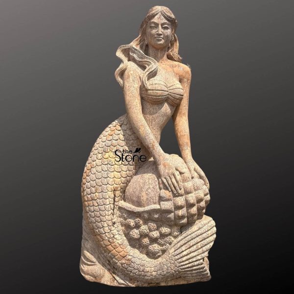 A striking 60-inch natural sandstone Mermaid Statue perfect for a garden, is presented from a frontal slightly elevated perspective against a plain dark background. The statue is carved from brown sandstone showcasing its natural texture and earthy tones which would beautifully complement an outdoor setting. The mermaid figure is depicted in a gracefully seated pose with her upper body upright and her tail curving beneath her. Her human-like upper torso features long, wavy hair cascading over her shoulders and a serene expression on her face. Her arms are positioned around a large, textured object in front of her resembling a cluster of fruit possibly grapes or pinecones within a basket-like base. Her scaled mermaid tail is prominently displayed, with individual scales intricately carved, and the fins at the end of her tail are also detailed. The entire sculpture exudes a classical mythical charm ideal for a focal point in a garden. The natural variations in the sandstone texture enhance its organic appeal. This detailed image vividly conveys the artistic beauty and natural aesthetic of the sandstone mermaid statue making it highly relevant to an article discussing natural stone mermaid statues for gardens.