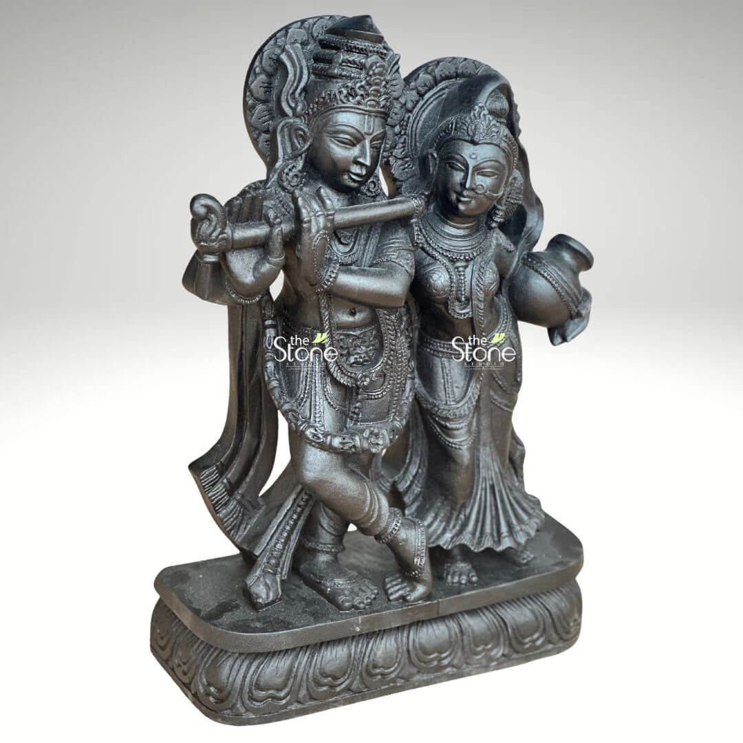 2ft Radha Krishna Black Stone Statue 6 A magnificent 2-feet black stone Radha Krishna idol is depicted in a slightly elevated frontal shot against a plain light background. The statue exquisitely portrays Lord Krishna and Radha standing together on an ornate rectangular base adorned with a lotus petal design. Lord Krishna on the left is captured in his iconic posture playing the flute held delicately to his lips with one leg crossed gracefully. He is richly embellished with an elaborate crown, intricate necklaces armlets and flowing dhoti all finely carved into the dark stone. Radha stands beside him on the right, her face gently turned towards Krishna embodying profound devotion. She carries a traditional pot or kalash in her left hand and is equally adorned with a crown, detailed jewelry and gracefully draped attire. Both figures are sculpted from deep black stone which imparts a timeless and spiritual presence with the fine details highlighted by ambient lighting. A subtle halo or decorative arch behind Krishna's head further enhances the divine composition. This detailed image vividly conveys the spiritual essence and artistic craftsmanship of the 2-foot black stone Radha Krishna statue making it highly relevant to an article on such idols.