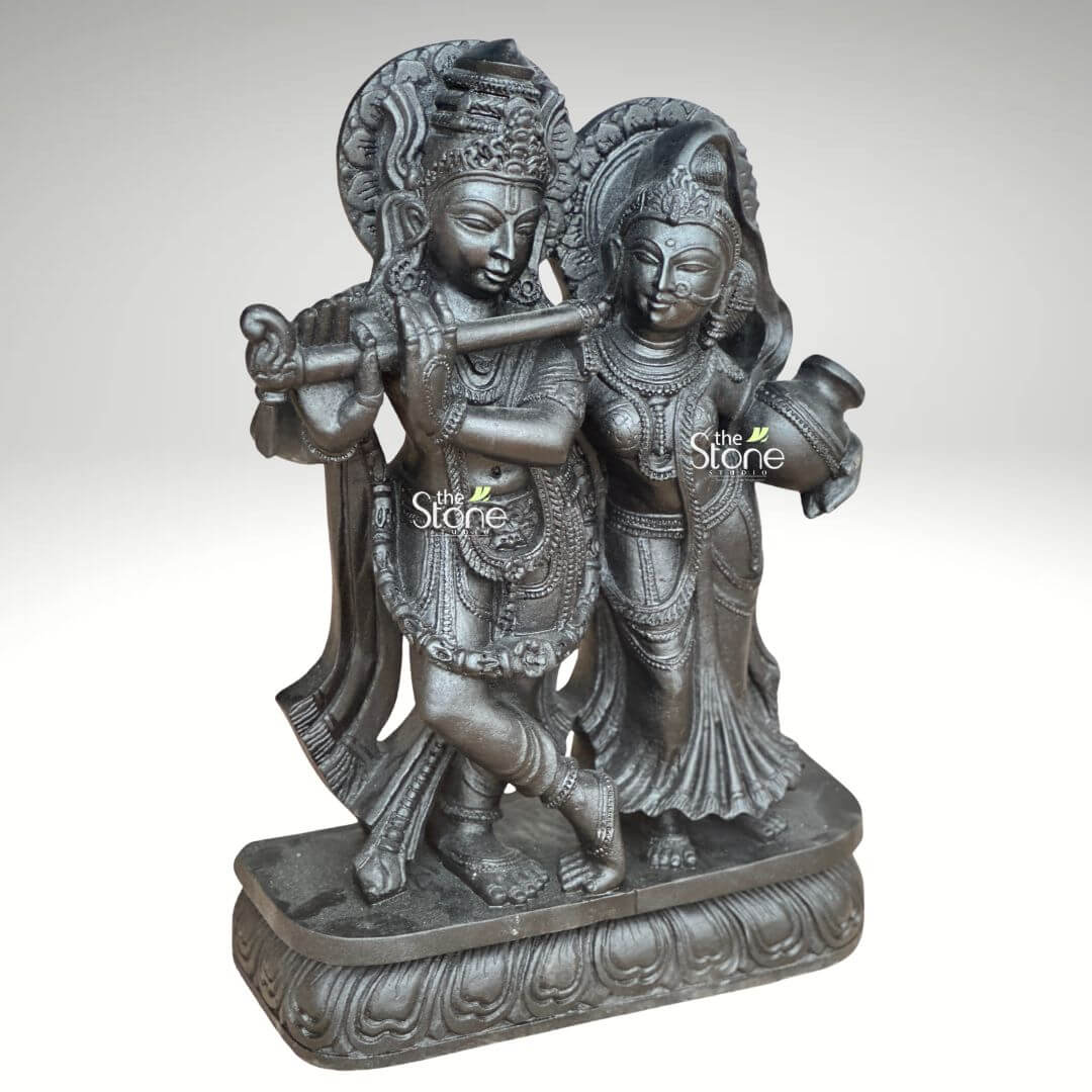 2ft Radha Krishna Black Stone Statue 4 An expertly hand-carved 2-foot tall black stone Radha Krishna sculpture masterfully designed to serve as a spiritual centerpiece for a home mandir or office. Shown from a three-quarter side angle to highlight its depth and volume the 2ft Radha Krishna statue captures the divine couple in a traditional standing posture. Lord Krishna is depicted in his iconic cross-legged Tribhanga pose gracefully playing a stone flute with a serene facial expression. Standing elegantly beside him is Goddess Radha looking toward him with a gentle and compassionate expression while holding a sacred water pot in her left hand. Both figures are adorned with towering tiered crowns mukuts intricate layered necklaces and armlets that showcase the exceptional craftsmanship of traditional Indian stone masonry. The sculpture features highly detailed relief work including grand circular Prabhavalis with rhythmic patterns framing their heads and realistic fluid fabric folds in their traditional attire. The entire black stone idol rests on a wide, multi-tiered pedestal embellished with delicate lotus-petal relief carvings. The deep charcoal-black hue and matte rustic finish of the natural stone provide a timeless temple-inspired aesthetic making it an ideal piece to invite peace prosperity and divine love into any modern or traditional living environment.