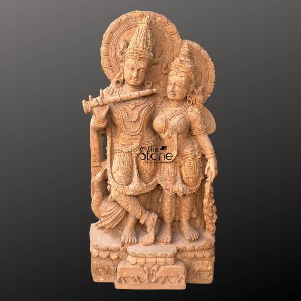 An exquisitely hand-carved Radha Krishna sandstone garden statue masterfully sculpted from a single block of natural brown sandstone with a rich grainy texture. The sculpture depicts the divine couple Lord Krishna and Goddess Radha in a traditional standing pose. On the left Krishna is shown in his iconic Tribhanga posture with his legs crossed gracefully holding a flute to his lips. Radha stands elegantly to his right looking affectionately toward him. Both figures are adorned with intricate tiered crowns layered jewelry and circular haloes or Prabhavalis featuring delicate floral relief work. At their feet a small detailed cow rests symbolizing Krishna’s role as the divine cowherd. The entire statue is mounted on a sturdy rectangular pedestal carved with cattle and lotus motifs. The warm earthy reddish-brown tones of the natural sandstone give the piece a timeless antique aesthetic making it an ideal spiritual centerpiece for an outdoor garden temple sanctuary or meditative landscape to invite peace and divine energy.