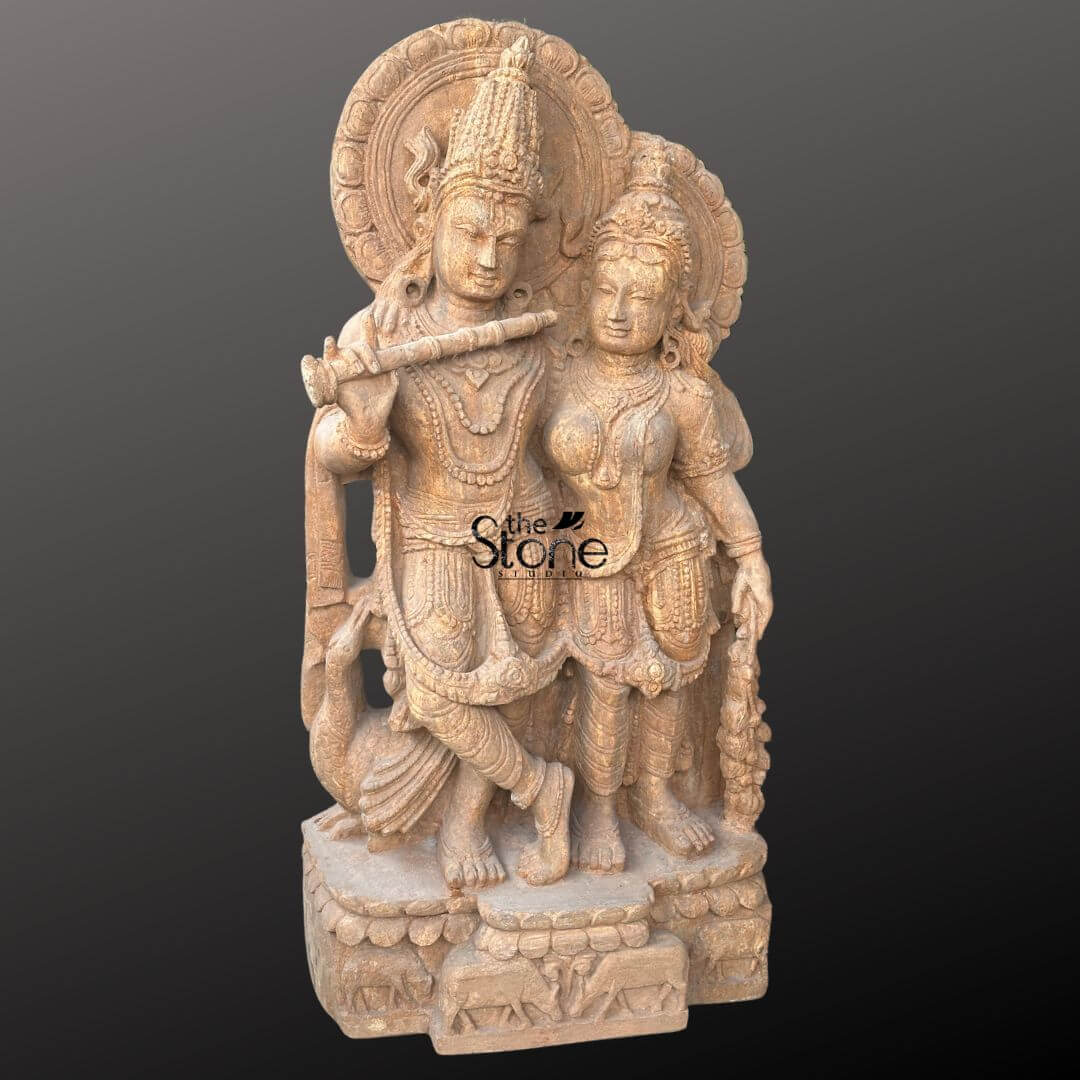 Radha Krishna Sandstone Garden Statue 3ft 5 An exquisitely hand-carved Radha Krishna sandstone statue masterfully sculpted from a single block of natural brown sandstone with a rich grainy texture. The sculpture depicts the divine couple in a traditional standing pose making it an ideal spiritual centerpiece for a home temple or mandir. On the left, Lord Krishna is shown in his iconic Tribhanga posture with his legs crossed, gracefully holding a stone flute to his lips. Standing elegantly to his right Goddess Radha looks toward him with a serene and gentle expression. Both figures are adorned with towering tiered crowns mukuts intricate layered necklaces, armlets, and circular Prabhavalis (haloes) featuring rhythmic floral and sunburst relief patterns. The craftsmanship is evident in the realistic fluid folds of their traditional attire and the delicate carving of a peacock standing loyally at Krishna’s feet. The entire sandstone Radha Krishna idol rests on a sturdy rectangular pedestal embellished with relief carvings of cattle and lotus motifs, symbolizing the life of the divine cowherd. The warm, earthy reddish-brown tones of the natural stone provide a timeless antique temple aesthetic designed to invite an aura of peace, prosperity, and divine love into any residential sanctuary or garden landscape.