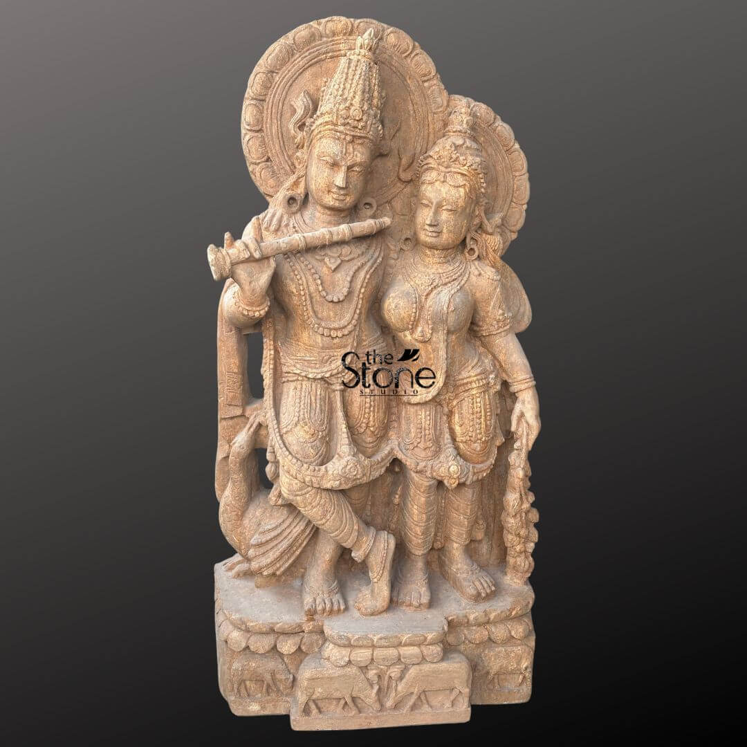 Radha Krishna Sandstone Garden Statue 3ft 6 An exquisitely hand-carved sandstone Radha Krishna statue masterfully sculpted from a single block of natural brown stone with a rich grainy texture. The sculpture depicts the divine couple Lord Krishna and Goddess Radha in a traditional standing pose that radiates serenity and grace. On the left Krishna is shown in his iconic Tribhanga posture with his legs crossed at the ankles, gracefully holding a stone flute to his lips. Radha stands elegantly to his right, her gentle expression reflecting divine love and devotion. Both figures are adorned with towering, multi-tiered crowns intricate layered necklaces, armlets, and circular Prabhavalis featuring delicate floral and sunburst relief patterns. The craftsmanship is evident in the realistic fluid folds of their traditional attire and the delicate carving of a peacock standing loyally at Krishna’s feet. The entire sandstone Radha Krishna idol rests on a sturdy rectangular pedestal embellished with relief carvings of cattle and lotus motifs, symbolizing the life of the divine cowherd. The warm, earthy reddish-brown tones and rustic finish of the natural sandstone give the piece a timeless, antique temple aesthetic designed to serve as a profound spiritual centerpiece for a home mandir, luxury garden landscape, or meditation sanctuary.