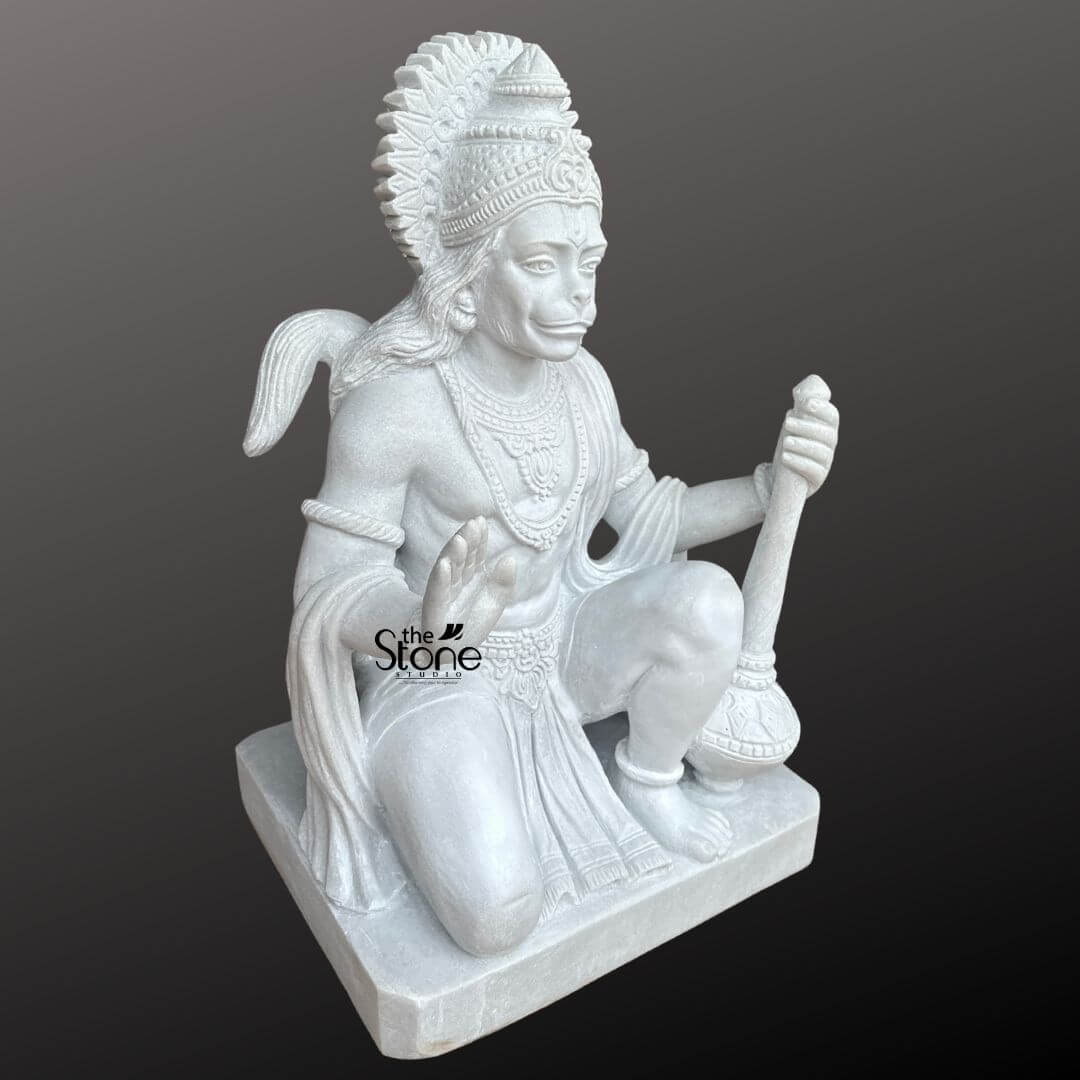 Hanuman Marble Statue for Home Temple 3ft 4 An exquisitely hand-carved 3-foot tall Sankat Mochan Hanuman idol masterfully sculpted from premium pure white marble with a smooth polished finish. The statue depicts Lord Hanuman in a powerful kneeling posture capturing his role as the legendary protector and remover of obstacles. His right hand is raised in the sacred Abhaya Mudra the universal gesture of fearlessness and blessing, while his left hand firmly holds his signature weapon the Gada positioned vertically on the base. The sculpture showcases exceptional artisan detail featuring a towering sunburst-style mukut layered necklaces, armlets, and the realistic, fluid fabric folds of his traditional dhoti. His facial expression is one of serene strength and profound devotion, complemented by a prominent white tilak. The entire 36-inch white marble Hanuman statue sits firmly on a solid square marble pedestal embodying stability and permanence. This large-scale spiritual icon serves as a majestic centerpiece for a home mandir temple sanctuary or meditation space designed to invite an aura of peace courage, and divine protection into the environment.
