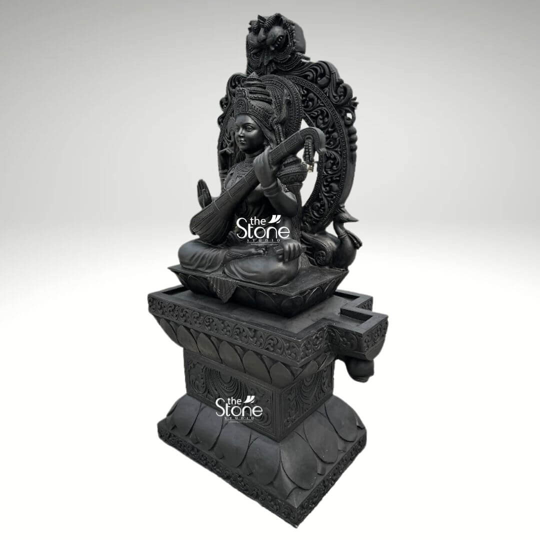 7ft Black Stone Saraswati Statue 9 A majestic 7-foot tall Saraswati statue masterfully hand-carved from premium natural black stone serves as a grand spiritual centerpiece for a temple foyer or a luxury home entrance. Shown from a side three-quarter perspective to highlight its monumental scale and intricate depth, the sculpture depicts the Goddess of knowledge, music, and the arts in a serene seated posture atop a massive, multi-tiered lotus pedestal. Lord Saraswati is portrayed with four arms, gracefully playing the Veena which symbolizes the harmony of all creative arts, while her upper hands hold a sacred book representing eternal knowledge and a mala for spiritual reflection. She is magnificently framed by a grand, circular Prabhavali featuring deeply carved traditional floral scrolls and a protective Kirtimukha face at its peak. The craftsmanship showcases incredible detail, from the towering tiered crown and large expressive eyes to the layered necklaces, armlets, and realistic fabric folds of her traditional attire. At the base of the statue, her divine vehicle, the swan is delicately rendered in relief. The refined matte charcoal finish of the stone highlights the heritage of traditional Indian stone masonry making this 7ft black stone Saraswati idol a powerful symbol of wisdom, purity, and creative inspiration designed to invite an aura of intellectual clarity and divine grace into any environment.