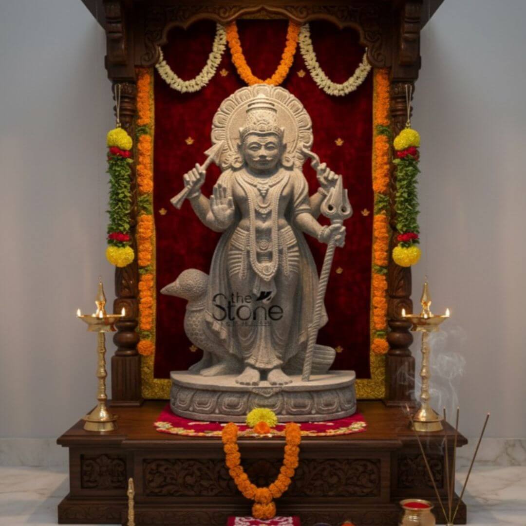 Shani Dev Stone Statue with Crow 27" 3 A serene and majestic home temple setup featuring a 27-inch Shani Bhagwan idol as the spiritual centerpiece. The Shani Dev statue masterfully hand-carved from natural blackstone with a rustic grey-matte finish, stands in a heroic posture atop an intricately detailed circular lotus pedestal. The deity is depicted with four arms, holding a traditional bow and arrow in his upper hands, with his lower right hand raised in the Abhaya Mudra (gesture of blessing and protection) and his left hand resting on a long, ornate staff. The blackstone Shani Dev sculpture is housed within an intricately carved dark wood mandir featuring ornate pillars and traditional motifs. The backdrop is a rich, deep red velvet fabric, beautifully framed by garlands of yellow marigolds and white blossoms. Standing faithfully behind the Lord is his divine vehicle, the crow carved with lifelike precision. In the foreground, the devotional atmosphere is enhanced by two tall, polished brass samai lamps with lit flames and wisps of smoke rising from burning incense sticks. The entire arrangement is set on a polished white marble floor, illustrating how a large-scale stone Shani Dev idol can be integrated into high-end residential interiors to invite discipline, stability, and divine protection into the home.