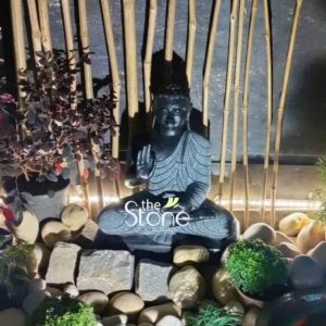 A serene 15-inch black stone Buddha statue ideal for a tranquil setting in Lucknow is beautifully arranged amidst natural elements. The Buddha is depicted in a seated meditative posture with his right hand raised in a gesture of fearlessness or blessing and his left hand resting on his lap. The black stone material gives the statue a profound almost mystical appearance with subtle details etched into the robes. The statue is surrounded by a carefully curated arrangement of smooth river stones and rocks of various sizes and shades creating a natural contemplative garden feel. Small green plants and moss-like clusters are interspersed among the stones, adding organic texture. In the background, tall bamboo stalks form a vertical screen casting long shadows and enhancing the peaceful ambiance. To the left, a potted plant with dark red foliage adds a touch of color. The lighting appears soft and atmospheric, highlighting the Buddha's serene expression with downcast eyes and the intricate patterns on his robe. This detailed image effectively conveys the spiritual essence and aesthetic appeal of the small black stone Buddha statue, making it highly relevant to an article discussing its presence and decor in Lucknow.