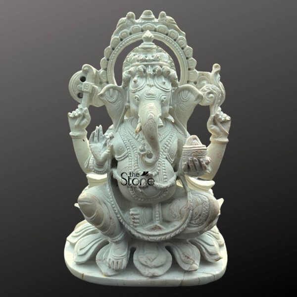 A finely crafted white stone Ganesha statue perfectly sized as a small idol for a car dashboard is featured against a plain dark background. The statue depicts Lord Ganesha in a traditional seated posture almost squatting upon a multi-layered lotus base symbolizing spiritual purity. He is shown with four arms his upper right hand holds an axe or goad and his upper left hand holds a noose (partially visible). His lower right hand is raised in a gesture of blessing while his lower left hand gently holds a small bowl of modaks, towards which his trunk gracefully curves. The pristine white stone material beautifully accentuates the intricate details of his elaborate crown jewelry and the folds of his dhoti. A decorative halo or prabhavali with a detailed outer edge frames Ganesha's head, enhancing his divine presence. The smooth finish and precise artistry of the carving make this compact Ganesha idol both auspicious and aesthetically pleasing. This detailed image clearly illustrates the sacred and decorative qualities of the white stone Ganesha statue making it highly relevant to an article discussing car dashboard placement.
