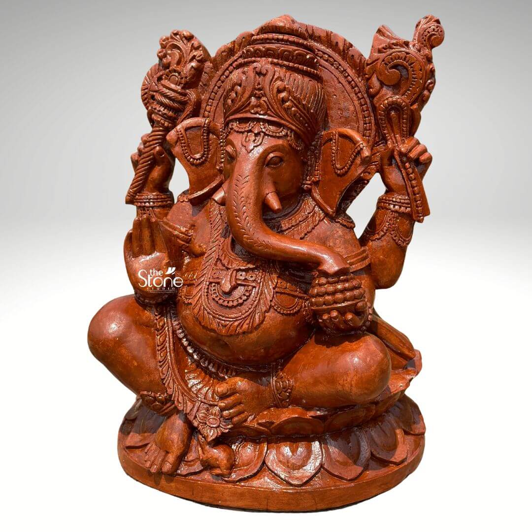 Pillaiyar Ganesha Statue 2.5ft 6 A magnificent 2.5-feet sandstone Sri Pillaiyar statue is presented from a frontal slightly elevated perspective against a plain light background. The statue is carved from reddish-brown sandstone showcasing its natural texture and warm, earthy tones. Lord Ganesha is depicted in a traditional, serene seated posture, cross-legged, on an intricately carved lotus base. He has four arms; his upper right hand holds an axe or goad, while his upper left hand grasps a noose. His lower right hand is raised in a gesture of blessing, and his lower left hand holds a bowl of modaks, with his trunk curving towards it. Ganesha is richly adorned with an elaborate crown, intricate necklaces, armlets, and a sacred thread draped across his plump belly, which symbolizes prosperity. His large, fan-like ears and expressive eyes are finely detailed. A magnificent halo or prabhavali, with a detailed outer edge and internal carvings, frames his head, emphasizing his divine stature. The textured surface of the sandstone highlights the intricate details of all the carvings, from the delicate features of Ganesha to the elaborate motifs on the backdrop and base. This detailed image powerfully conveys the spiritual majesty and artistic craftsmanship of the sandstone Sri Pillaiyar Ganesha statue, making it highly relevant to an article discussing such revered idols.
