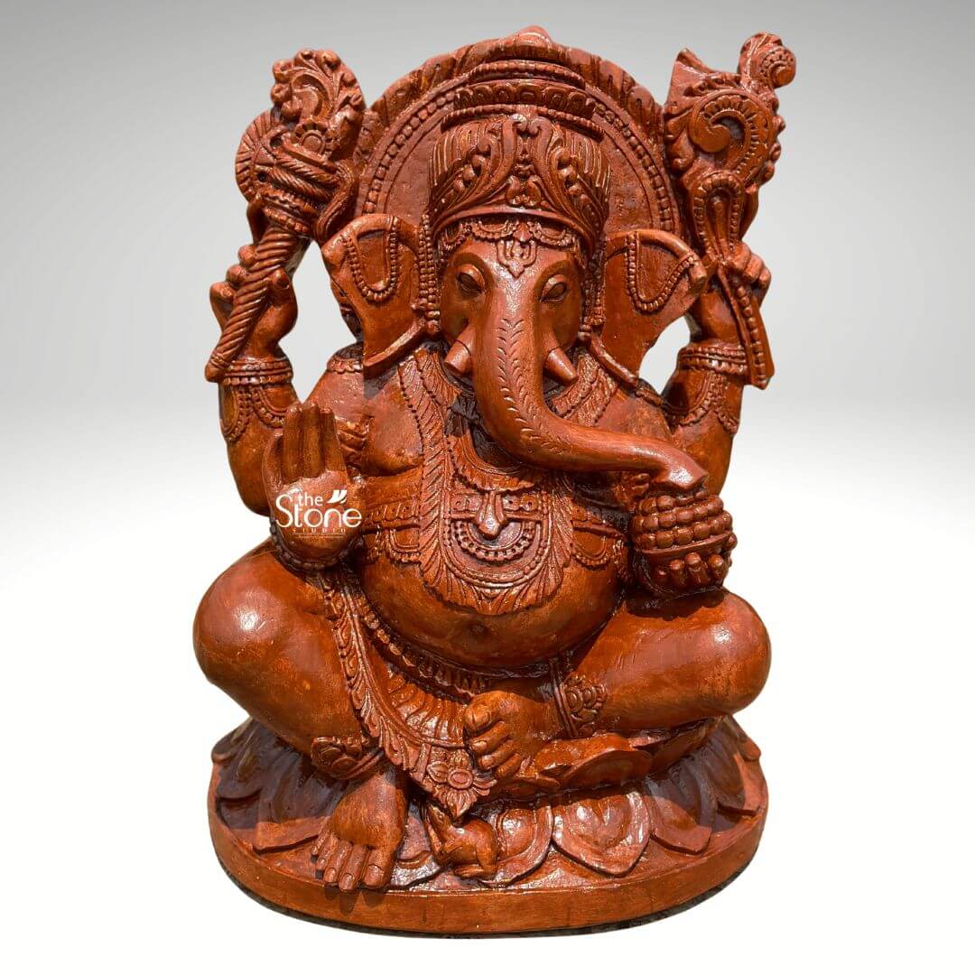Pillaiyar Ganesha Statue 2.5ft 5 A magnificent 2.5-feet sandstone Sri Vinayagar statue is presented from a frontal slightly elevated perspective against a plain light background. The statue is carved from reddish-brown sandstone, showcasing its natural texture and warm earthy tones. Lord Ganesha is depicted in a traditional serene seated posture, cross-legged, on an intricately carved lotus base. He has four arms; his upper right hand holds an axe or goad, while his upper left hand grasps a noose. His lower right hand is raised in a gesture of blessing and his lower left hand holds a bowl of modaks with his trunk curving towards it. Ganesha is richly adorned with an elaborate crown, intricate necklaces, armlets, and a sacred thread draped across his plump belly, which symbolizes prosperity. His large, fan-like ears and expressive eyes are finely detailed. A magnificent halo or prabhavali, with a detailed outer edge and internal carvings, frames his head, emphasizing his divine stature. The textured surface of the sandstone highlights the intricate details of all the carvings, from the delicate features of Ganesha to the elaborate motifs on the backdrop and base. This detailed image powerfully conveys the spiritual majesty and artistic craftsmanship of the sandstone Sri Vinayagar statue, making it highly relevant to an article discussing such revered idols.