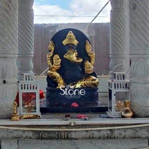 A striking 2-foot black stone Ganesha statue a significant piece in Bangalore, is prominently displayed within an outdoor temple-like setting possibly on a rooftop or terrace, under a bright sky. The Ganesha statue is carved from dark black stone, with its key features and attributes magnificently highlighted in gold, creating a stunning contrast. Lord Ganesha is depicted in a traditional seated posture, with his trunk curving towards his left. His hands and elements of his crown are gilded in shimmering gold. The black stone body serves as a deep rich background for the golden accents, emphasizing his divine form. The statue is placed on a simple dark platform, and in front of it, small pink flower petals and devotional items are scattered. The setting is framed by two ornate white stone pillars on either side, adding a sense of grandeur. Between the Ganesha and the pillars, two white decorative lanterns are placed; the left one holds a small lit oil lamp (diya), and the right one contains a golden vase. A traditional brass lamp and other small items are also visible. The background shows glimpses of a modern building and an open sky, suggesting an urban context. This detailed image powerfully conveys the spiritual majesty and artistic craftsmanship of the black stone Ganesha statue with golden embellishments, making it highly relevant to an article discussing its presence in Bangalore.