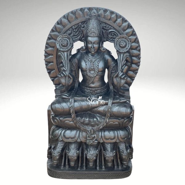 A majestic 2.5-feet black stone Surya Dev statue is presented from a frontal slightly elevated perspective against a plain light background. The sculpture depicts Surya Dev the Hindu solar deity seated in a regal posture embodying power and enlightenment. The entire statue is meticulously carved from rich dark black stone which gives it a profound and traditional presence, with a subtle, antique-like sheen. Surya Dev is shown with two arms each holding a blooming lotus flower symbols of purity and creation. He is richly adorned with an elaborate crown intricate necklaces armlets, earrings and a sacred thread across his chest. His serene face with slightly downcast eyes radiates benevolence. Behind his head rises a grand, circular prabhavali intricately carved with lotus petals radiating outwards symbolizing the sun's rays and his divine effulgence. The most distinctive feature of the statue is its base: Surya Dev is seated atop a chariot driven by seven horses, which are exquisitely carved in a row beneath his lotus throne representing the seven colors of the rainbow or the seven days of the week. The smooth yet detailed finish of the black stone highlights the fine artistry and the powerful benevolent presence of the solar deity. This detailed image powerfully conveys the spiritual majesty and artistic craftsmanship of the black stone Surya Dev statue, making it highly relevant to an article discussing such revered idols.