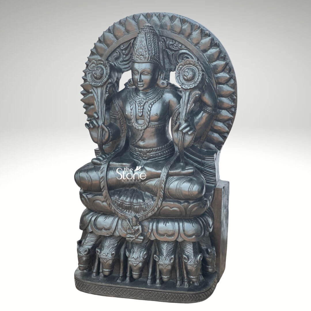 Surya Dev Black Stone Statue 30" 7 An exquisitely hand-carved **2.5-foot tall Surya Dev Murti**, masterfully sculpted from premium **natural black stone** with a refined matte charcoal finish. This spiritual masterpiece depicts the Sun God in a traditional seated posture atop a majestic, multi-tiered pedestal, radiating an aura of cosmic discipline and divine energy. **Lord Surya** is portrayed with two arms, each holding a long-stemmed, blooming lotus flower, which symbolizes purity and the awakening of consciousness. The deity is adorned with a towering, tiered **mukut (crown)** and intricate temple jewelry, including layered necklaces and armlets that showcase the exceptional craftsmanship of traditional Indian stone masonry. Framing the head and upper body is a grand, circular **Prabhavali (sunburst halo)** embellished with rhythmic, petal-like relief patterns that represent the life-giving rays of the sun. A defining iconographic feature of this **black stone Surya Dev statue** is the **seven horses (Saptashva)** carved in high relief at the base of his celestial chariot, representing the seven days of the week and the seven colors of light. This durable and weather-resistant **30-inch stone sculpture** serves as a profound spiritual icon, ideal for a home mandir, grand foyer, or office space, designed to invite vitality, success, and positive energy into any environment.