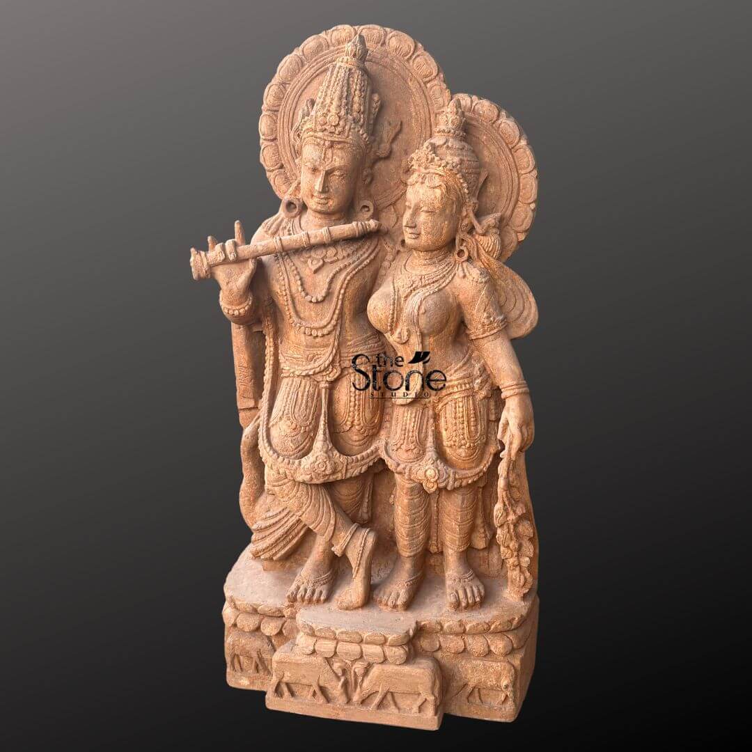Radha Krishna Sandstone Garden Statue 3ft 7 An exquisitely hand-carved traditional Radha Krishna sandstone idol masterfully sculpted from a single block of natural brown sandstone with a rich grainy texture. This spiritual masterpiece depicts the divine couple Lord Krishna and Goddess Radha in a traditional standing pose that radiates serenity and grace. On the left Lord Krishna is shown in his iconic Tribhanga posture with his legs crossed at the ankles gracefully holding a stone flute to his lips. Radha stands elegantly to his right her gentle expression reflecting divine love and devotion. Both figures are adorned with towering multi-tiered crowns mukuts intricate layered necklaces armlets, and circular Prabhavalis (ornate haloes) featuring delicate floral and rhythmic relief patterns. The craftsmanship is evident in the realistic fluid folds of their traditional attire and the delicate carving of a peacock and cow standing loyally at the base. The entire sandstone Radha Krishna statue rests on a sturdy rectangular pedestal embellished with relief carvings of cattle and lotus motifs, symbolizing the life of the divine cowherd. The warm, earthy reddish-brown tones of the natural stone provide a timeless antique temple aesthetic designed to serve as a profound spiritual centerpiece for a home mandir, luxury garden landscape, or meditation sanctuary.