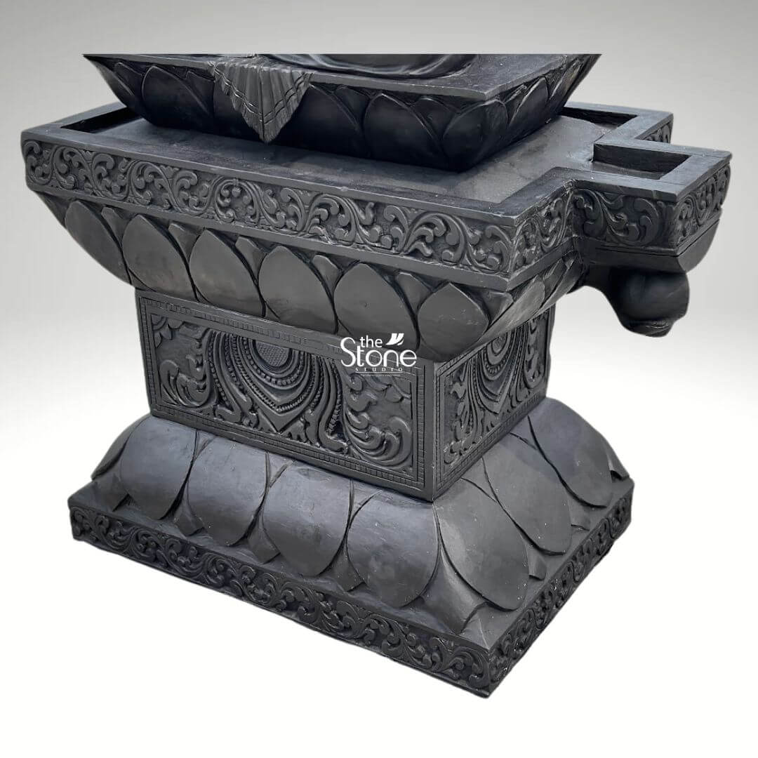 7ft Black Stone Saraswati Statue 11 A robust and elaborately carved black stone pedestal, clearly designed as a base for a large, traditional temple statue, is presented from a frontal, slightly elevated perspective against a plain light background. The pedestal is constructed in multiple tiers, all carved from dark black stone, showcasing exquisite traditional craftsmanship. The uppermost section features a wide, rectangular platform, with a central, stylized lotus design that appears to function as a deep oil lamp or offering bowl. This central feature is flanked by smaller, rectangular indentations, also for offerings or wicks. The main body of the pedestal is richly adorned with deeply carved floral and scrollwork patterns along its upper edges, and a prominent, repetitive lotus petal motif on its lower sections. The central front face of the lower rectangular tier displays a highly detailed circular motif, possibly a sun disc or sacred symbol, surrounded by more intricate scrollwork and auspicious patterns. The entire structure exhibits a textured, matte finish typical of finely carved stone, suggesting both durability and sacred artistry. This detailed image focuses solely on the ornate black stone pedestal, which, in the context of the article, is an integral part of a traditional temple statue of Goddess Saraswati.