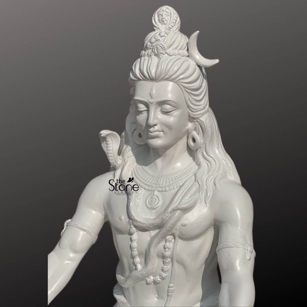 2ft Shiva Meditation Statue 5 A close-up slightly elevated shot captures the upper body of a 2-foot fiber white Lord Shiva statue ideal for home worship set against a plain dark background. The statue is entirely white emphasizing the divine purity and serenity of the deity. Lord Shiva's face is serene with gently closed or downcast eyes conveying deep meditation and inner calm. His intricately matted hair (Jata) is piled high, with a prominent crescent moon nestled within, and long, flowing locks cascade down his shoulders. A finely sculpted cobra is draped around his neck and shoulders, its head subtly resting near his left shoulder. He wears multiple strands of rudraksha beads as necklaces, and his powerful physique is subtly rendered. His third eye is marked on his forehead, and large, traditional earrings adorn his elongated earlobes. The smooth lustrous finish of the fiber material highlights every detail of his powerful yet tranquil form, from the muscular definition to the delicate curves of his features. This detailed image effectively conveys the spiritual essence and artistic craftsmanship of the fiber white Lord Shiva statue making it highly relevant to an article discussing home idols.