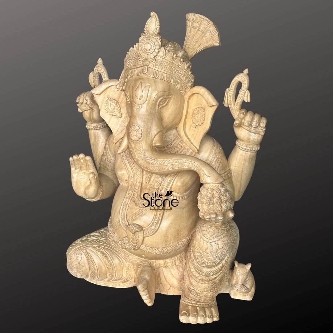 Wooden Ganesha Statue Online 2ft 6 A masterfully hand-carved 2-foot tall wooden Ganesh murti for home sculpted from natural light-toned wood with a smooth polished finish. This spiritual idol depicts Lord Ganesha in a stable seated posture showcasing a serene meditative expression with large expressive ears and a gracefully curved trunk reaching for a bowl of modaks held in his lower left hand. Lord Ganesha’s four arms carry traditional Vedic symbols while his lower right hand is raised in the Abhaya Mudra the universal gesture of protection and blessing. The sculpture features intricate detailing on the towering tiered crown layered necklaces and armbands along with realistic fabric folds on his traditional dhoti. At the base of the statue, a small figure of his loyal vehicle the mouse, is delicately carved near his foot. This 24-inch wooden Ganesha sculpture serves as an ideal centerpiece for a home mandir, foyer, or living room, blending traditional Indian craftsmanship with a warm, organic aesthetic to invite peace and prosperity into the living space.