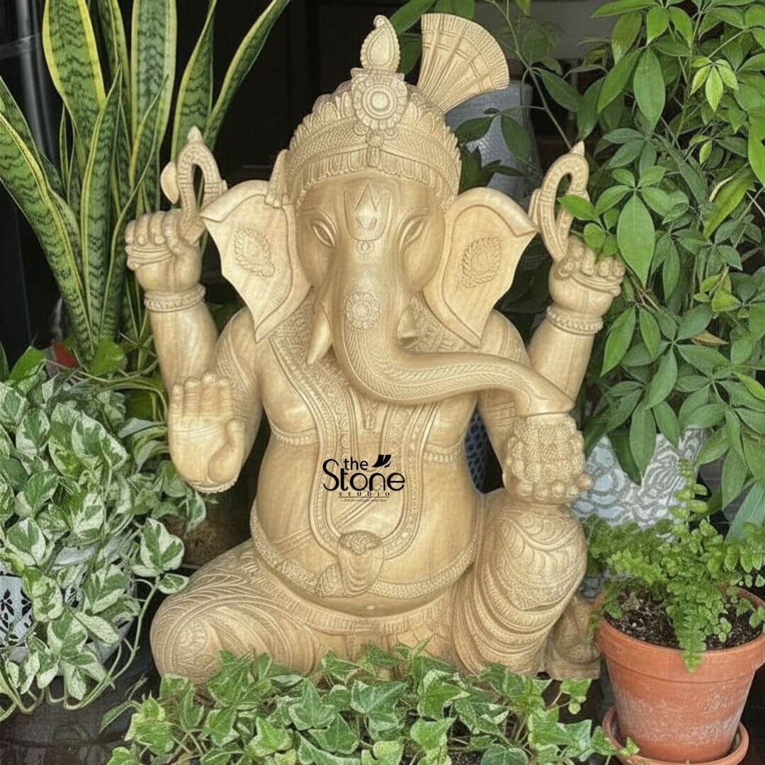 Wooden Ganesha Statue Online 2ft 3 A beautifully carved 2-foot wooden Ganesh Murti suitable for both temples and as a gift is positioned amidst lush green plants in an indoor or sheltered outdoor setting. The statue is crafted from light-colored wood showcasing its natural grain and warm inviting tones that blend harmoniously with the botanical surroundings. Lord Ganesha is depicted in a traditional seated posture with one knee raised exuding an aura of wisdom, prosperity, and benevolence. Ganesha is shown with four arms his upper hands hold traditional attributes possibly a noose and an axe. His lower right hand is raised in a gesture of blessing offering protection, while his lower left hand rests on his lap, possibly holding a modak. His elephant head is intricately detailed, with large, fan-like ears, expressive eyes, and his trunk curving gracefully towards his left. He wears an elaborate crown adorned with a fan-shaped element at the back, along with intricate necklaces, armlets, and a sacred thread across his plump belly. His garments are finely sculpted with delicate folds and patterns, enhancing the traditional aesthetic. The natural wood finish adds to its rustic and handcrafted charm. The surrounding plants include snake plants, pothos, ferns in terracotta pots, and other leafy greens, creating a serene and natural backdrop. This detailed image vividly conveys the spiritual majesty and artistic craftsmanship of the wooden Ganesha statue, making it highly relevant to an article discussing its use for temples or as a thoughtful gift.